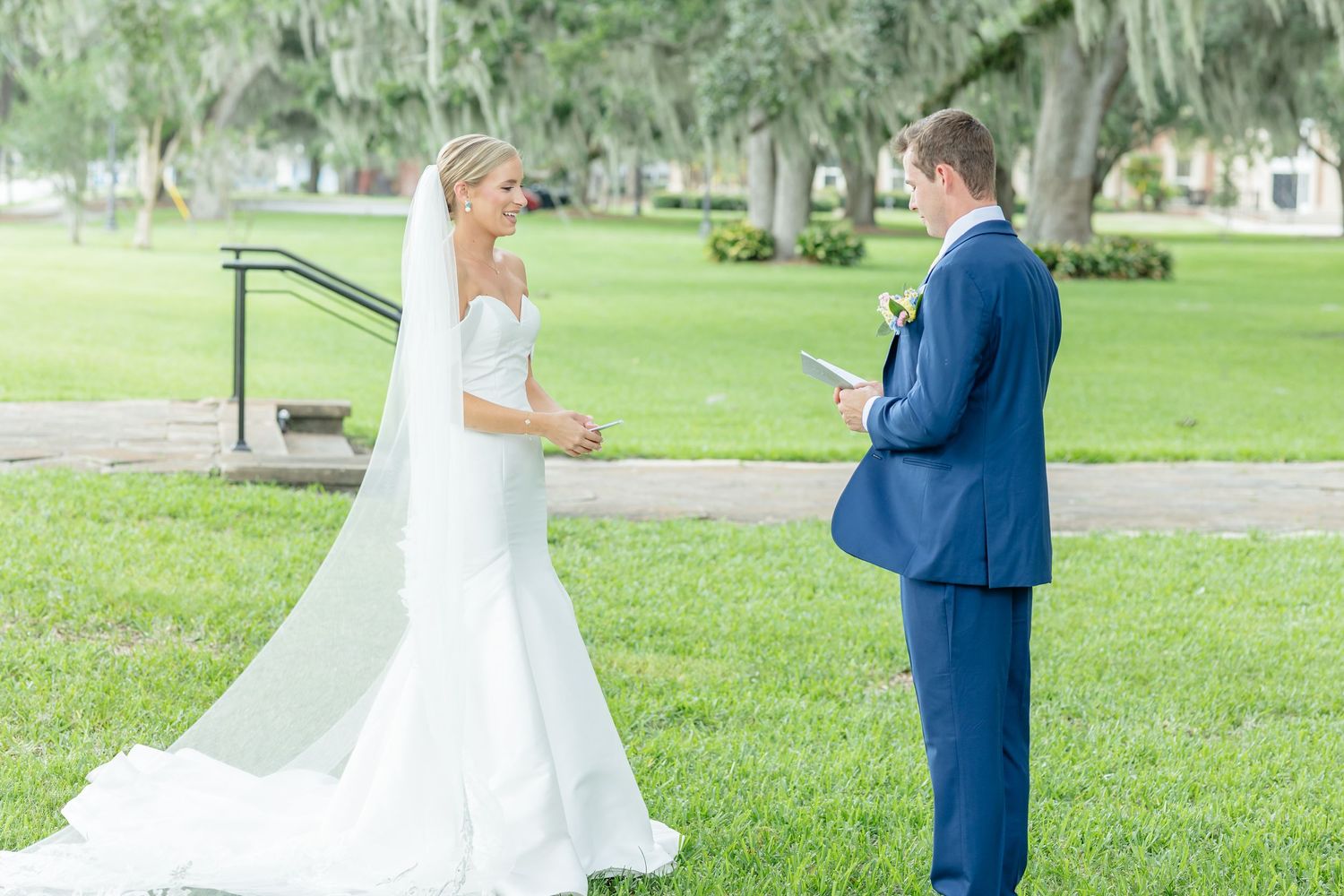 A couple in wedding attire stands together on a lush green lawn during an outdoor ceremony.