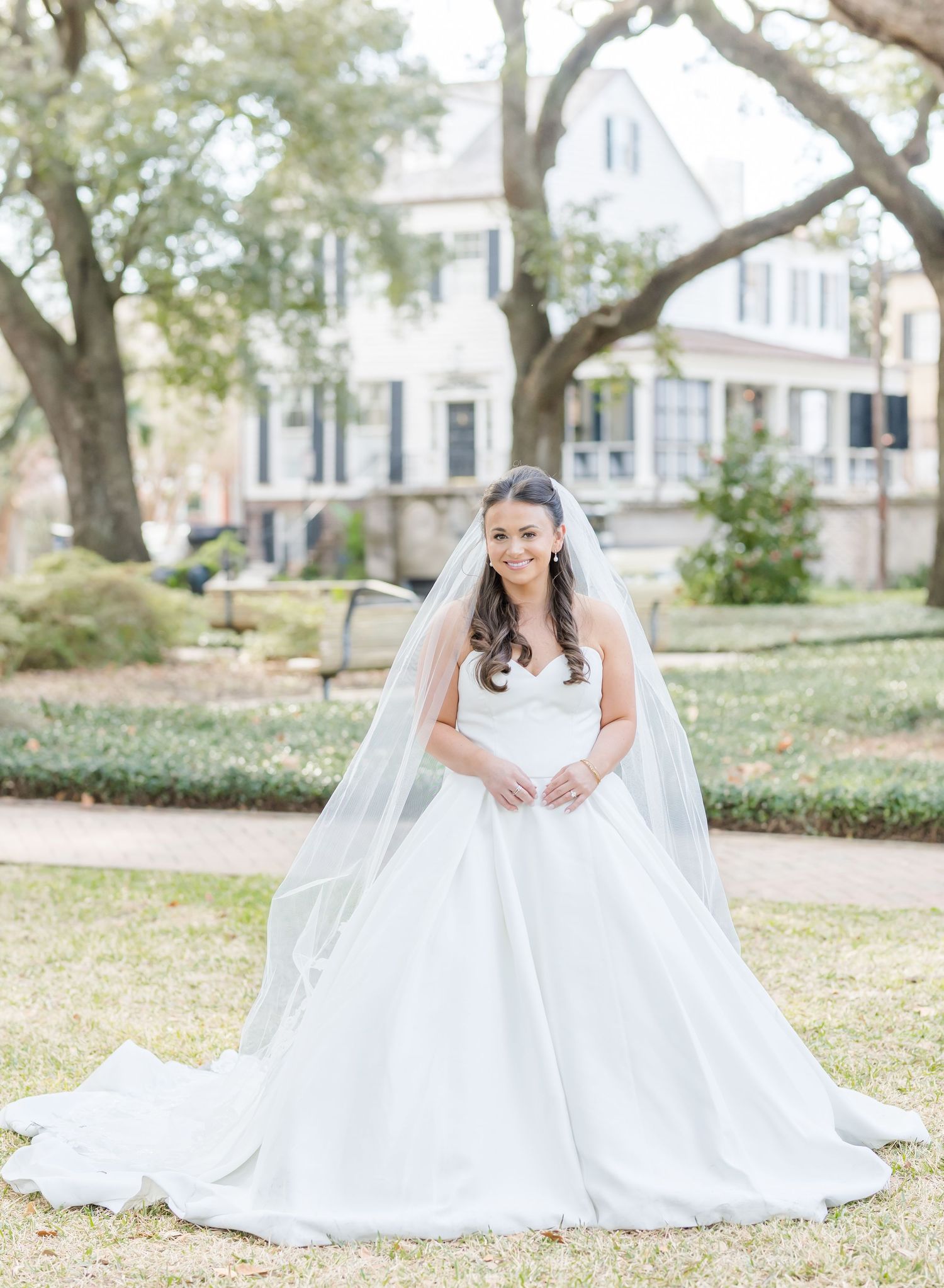 A bride in a flowing white ballgown poses elegantly on grass near historic buildings.