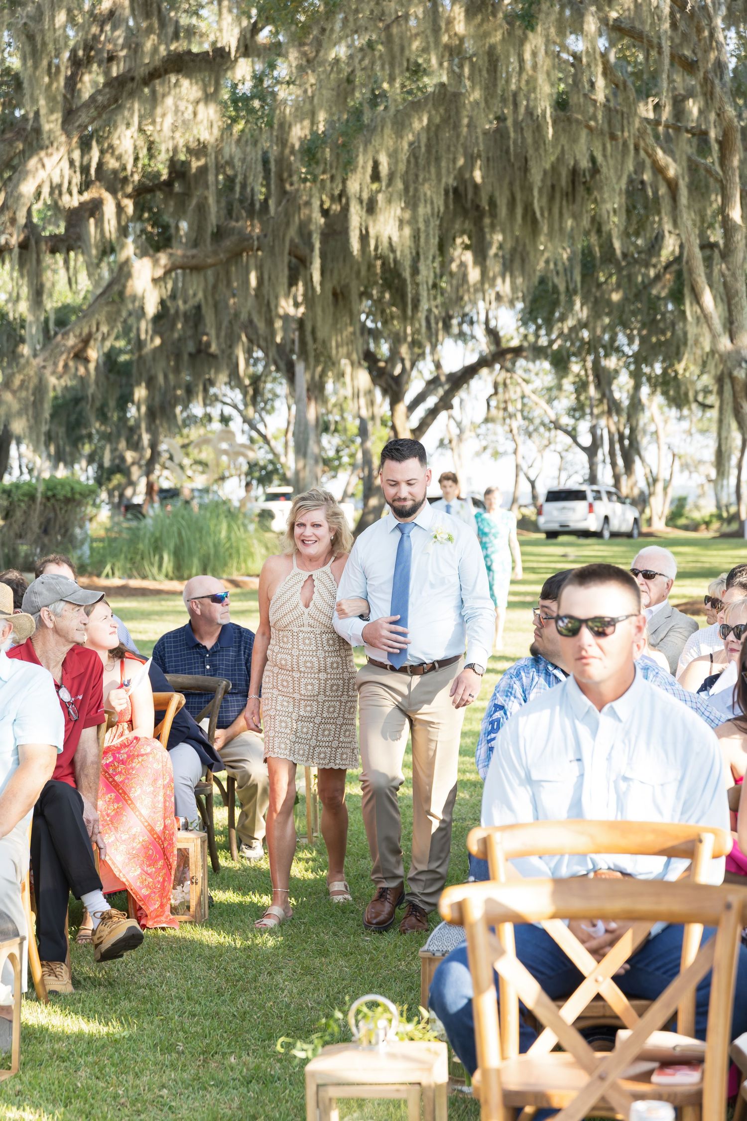 Outdoor gathering under moss-draped oak trees with wooden benches and people seated in a shaded area.