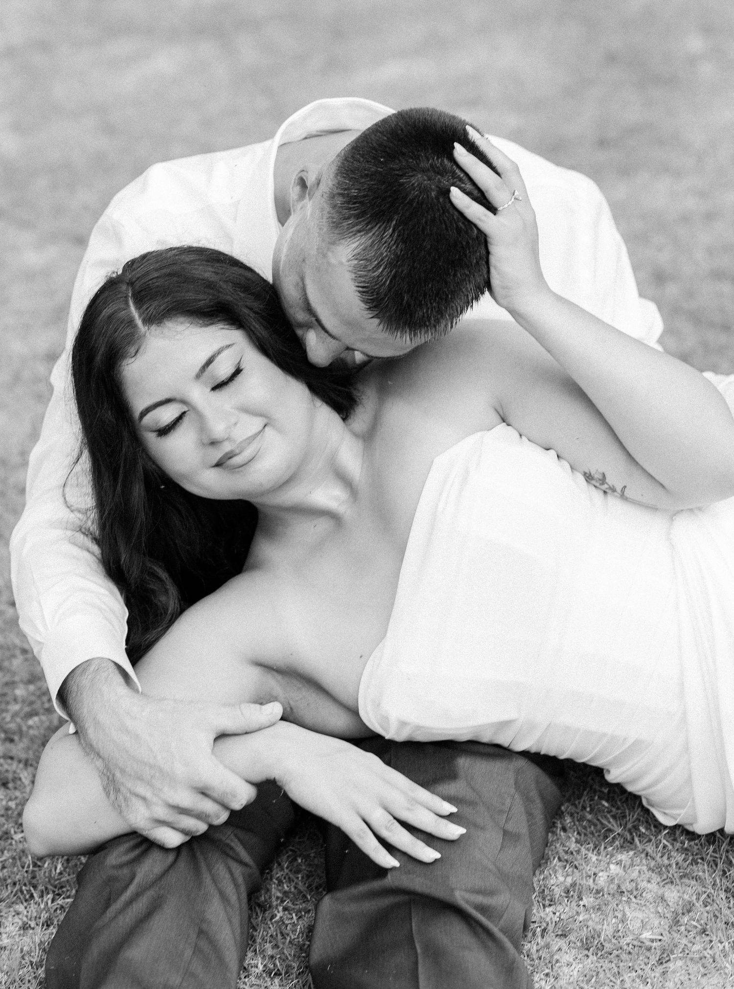 Black and white romantic photo of couple sitting on grass, man tenderly kissing woman's head.