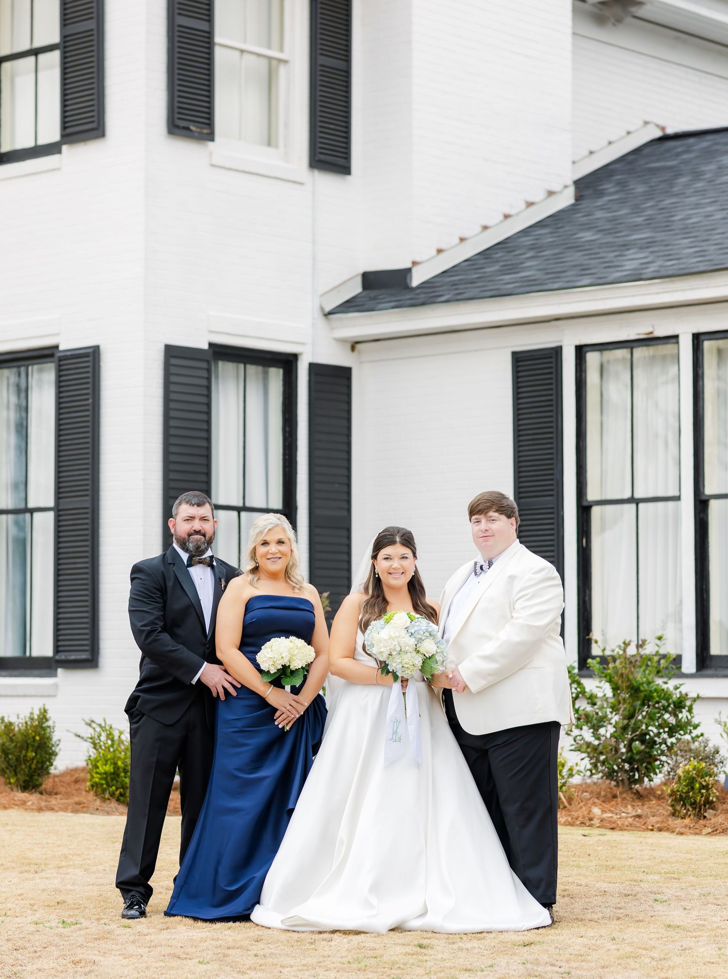 Bride and groom pose with two family members in navy formal wear outside a white building with black shutters.