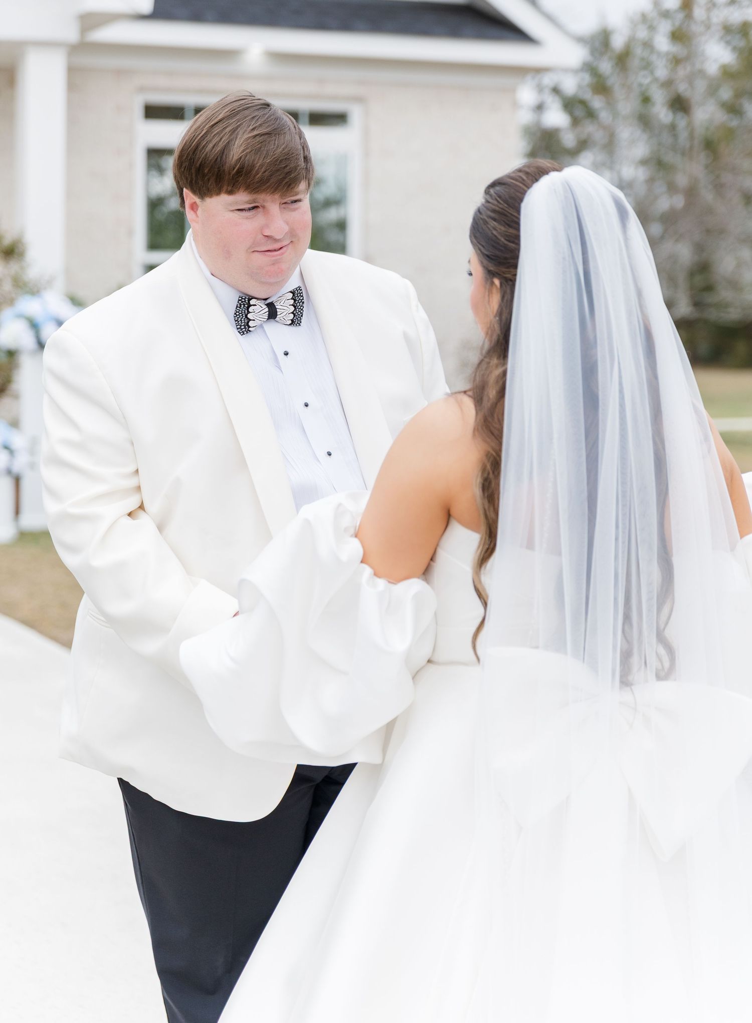 Groom in white tuxedo jacket holds bride's hands as she faces him in strapless gown and veil.