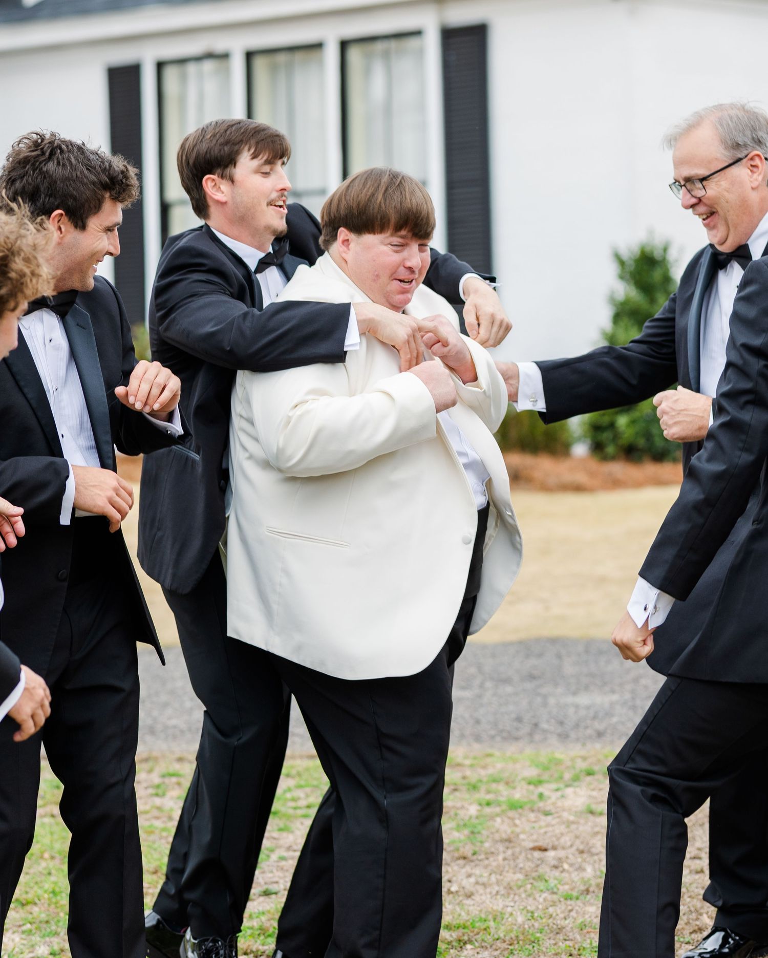 Groom in white jacket playfully wrestles with groomsmen in black tuxedos outside wedding venue.