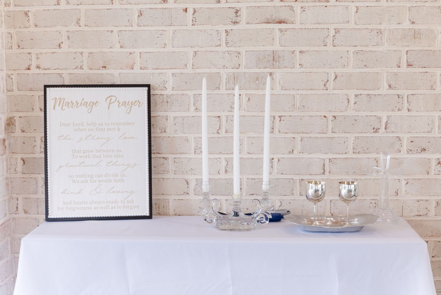 Wedding unity candle table with white taper candles and a framed Marriage Prayer sign against a whitewashed brick wall.