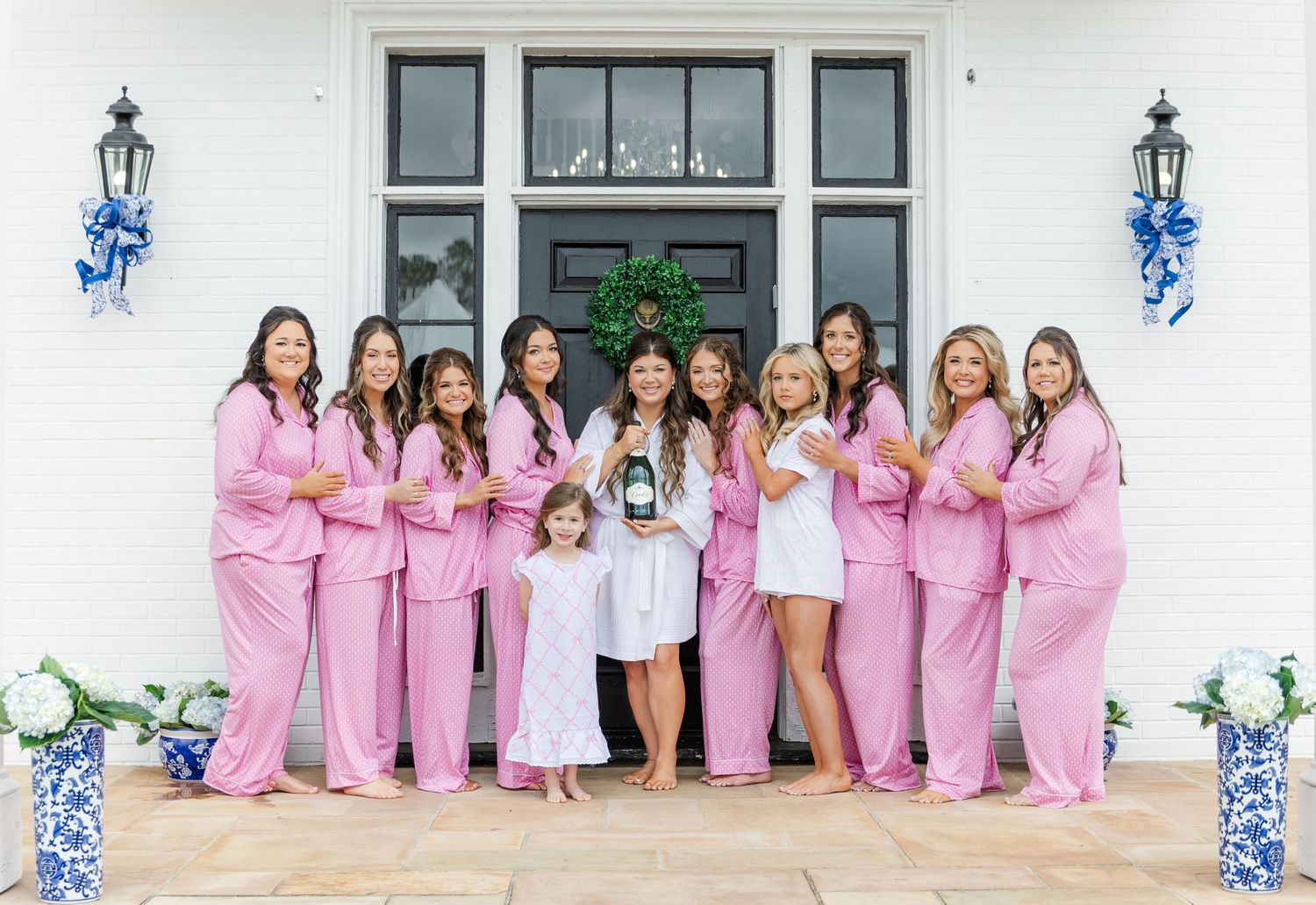 Bride in white robe with bridesmaids in matching pink pajamas posing on steps of a white wedding venue entrance.