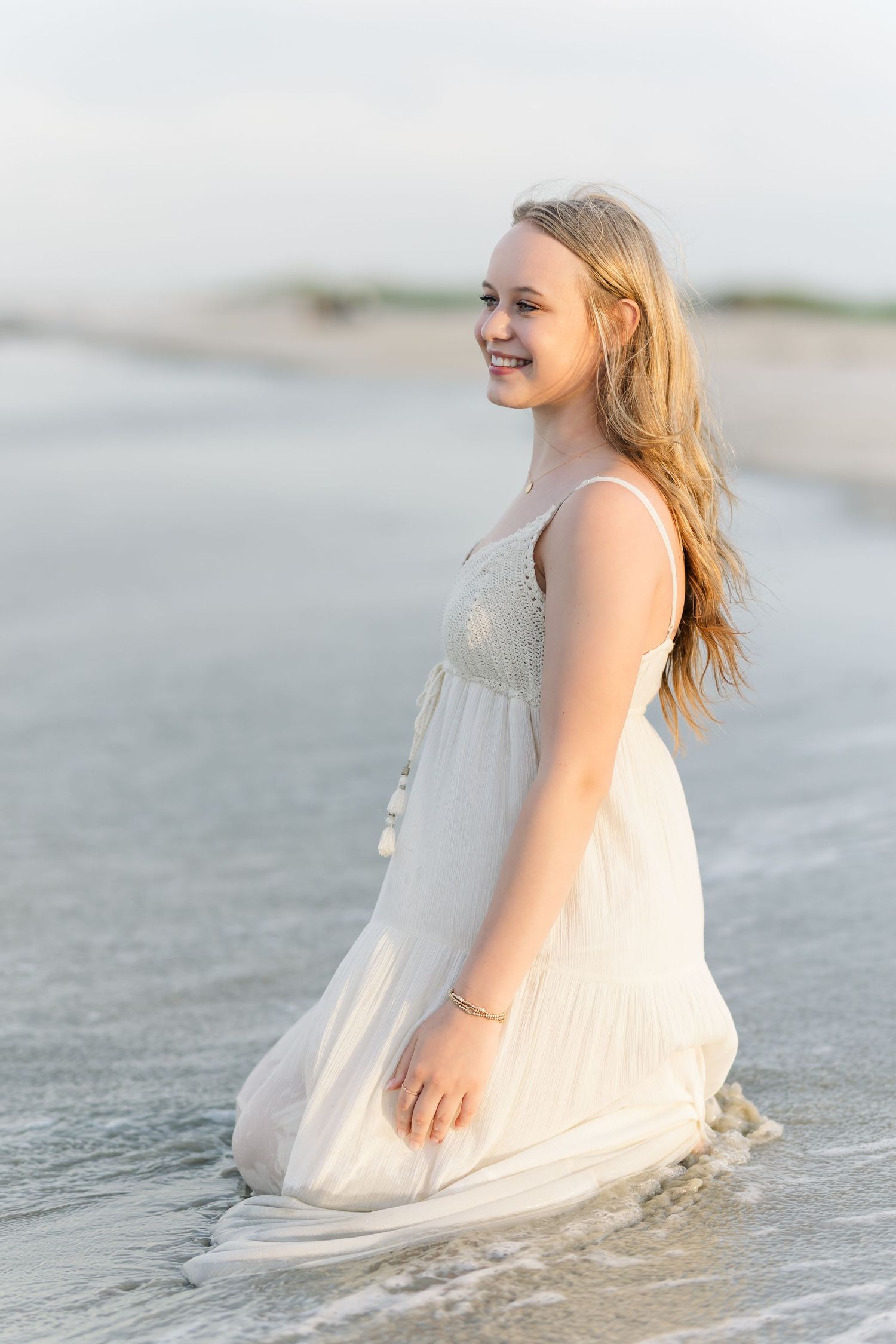 Candid moments captured sitting in gentle waves wearing a white summer dress at the shoreline.