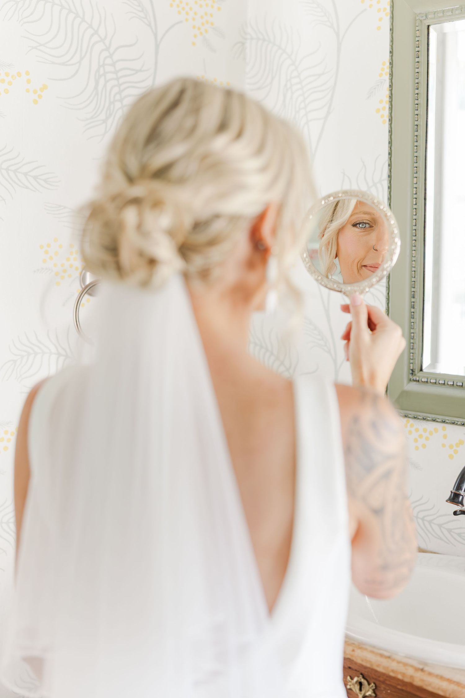 Blonde bride with tattoos checking makeup in hand mirror wearing white wedding gown and veil.