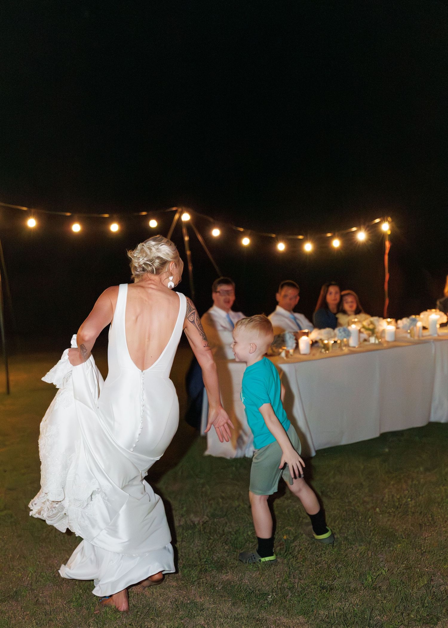 Bride in backless white gown dances with young boy under string lights at outdoor wedding reception.