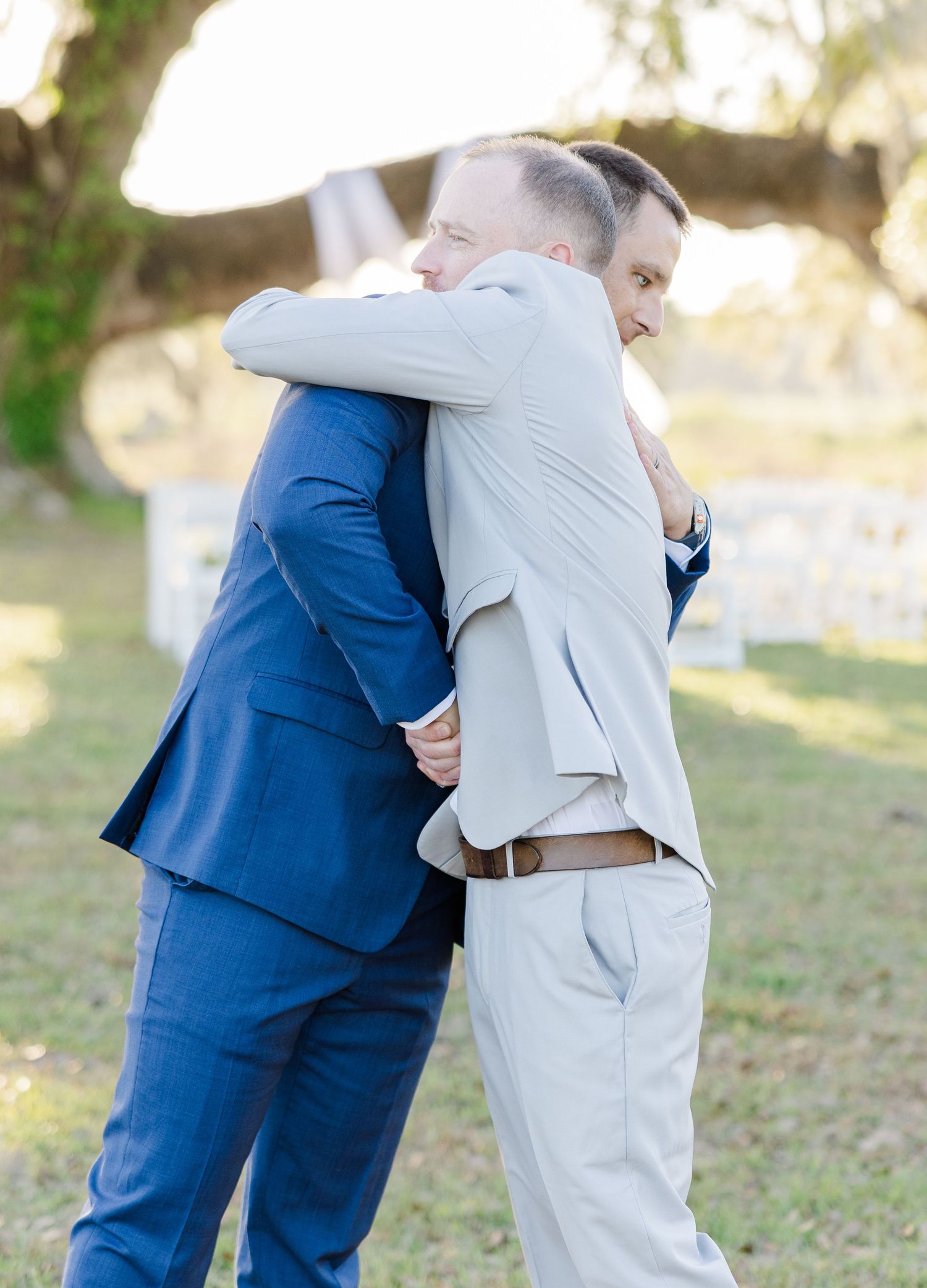 Two groomsmen sharing a heartfelt hug in suits before an outdoor wedding ceremony.