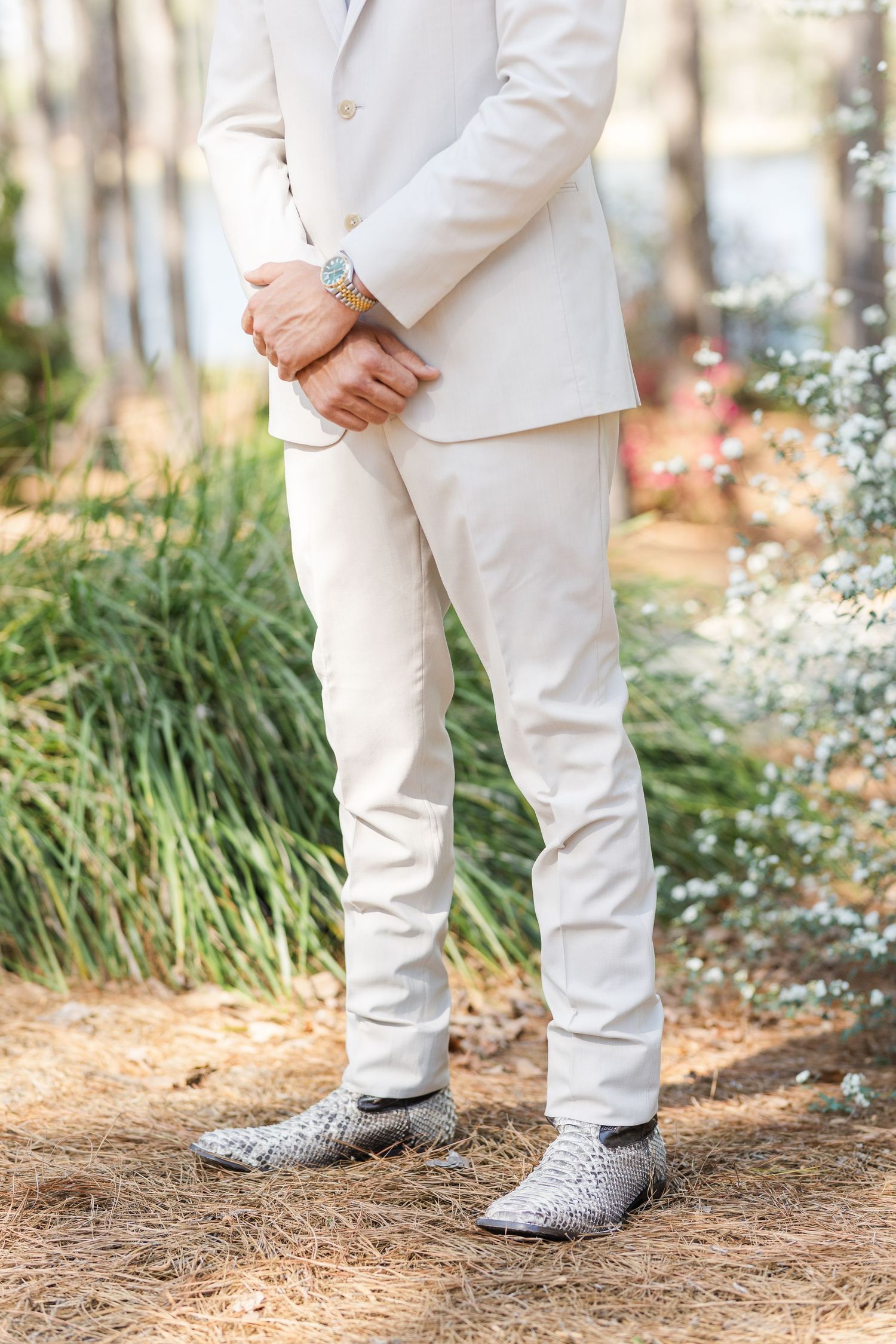 Groom in ivory suit with silver snakeskin dress shoes standing outdoors among pine trees.