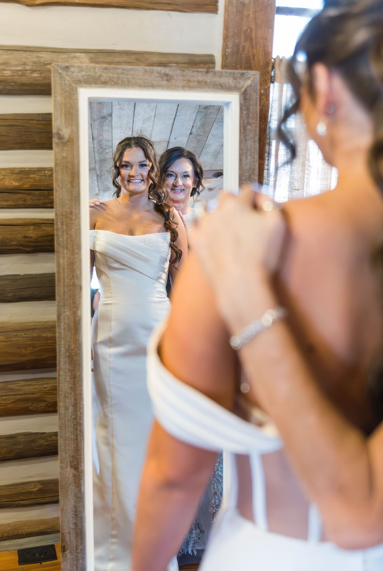 Bride and bridesmaid smiling together in a rustic wood-framed mirror while bride adjusts her earring.