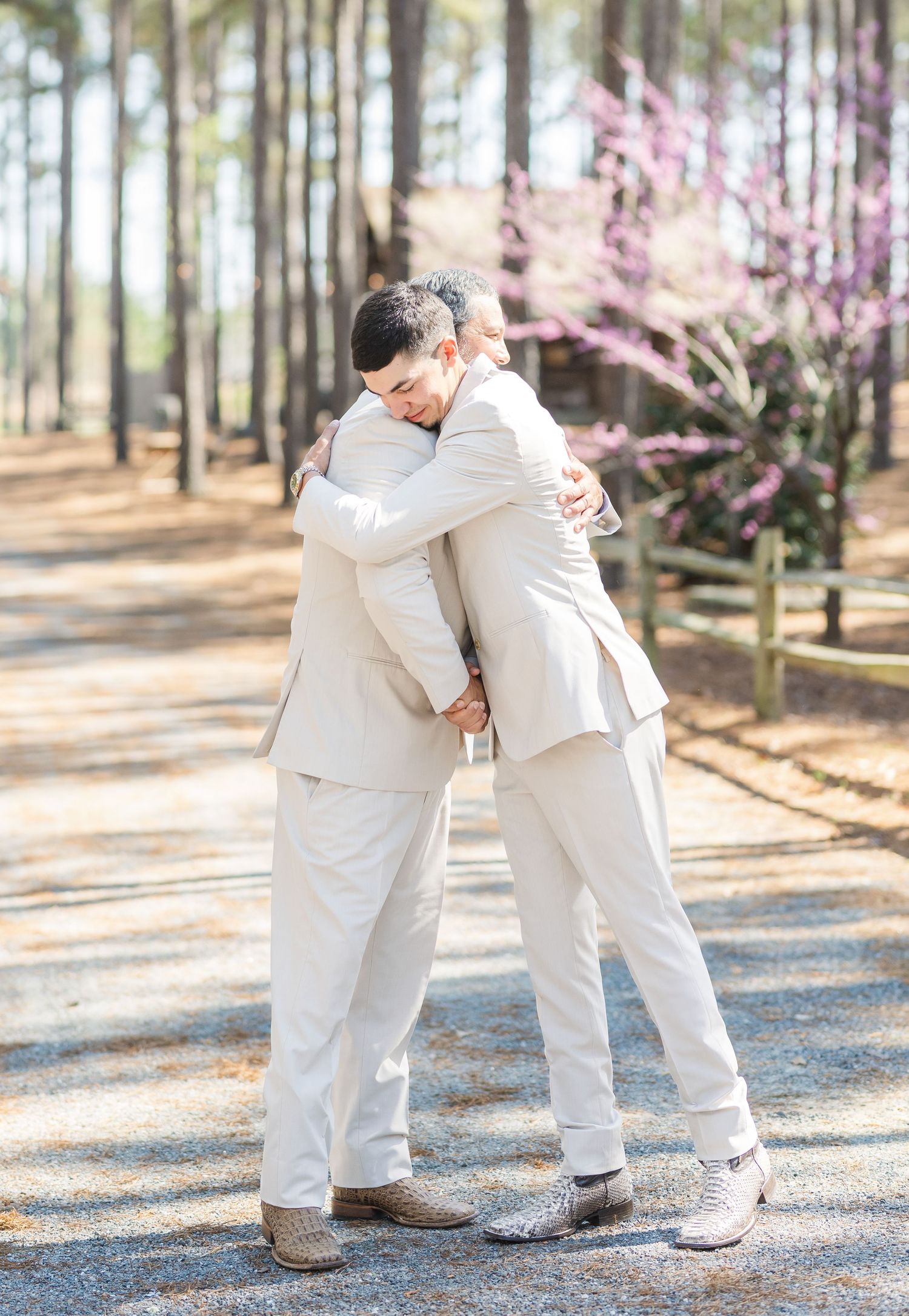 Two groomsmen in matching cream suits share an emotional hug outdoors at a spring wedding ceremony.