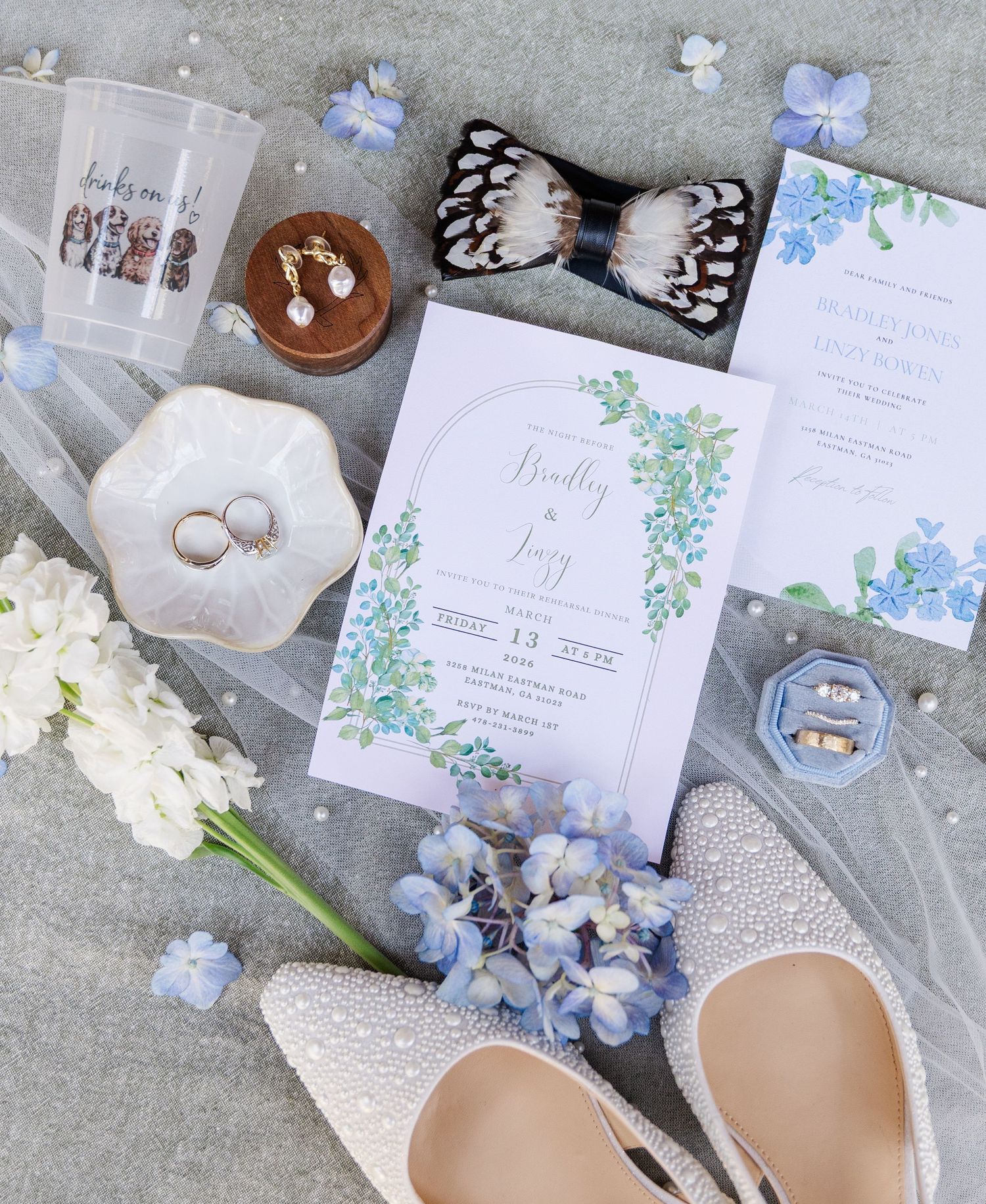 Blue hydrangea wedding flat lay with invitation, pearl heels, rings, feather bow tie, and floral stationery.