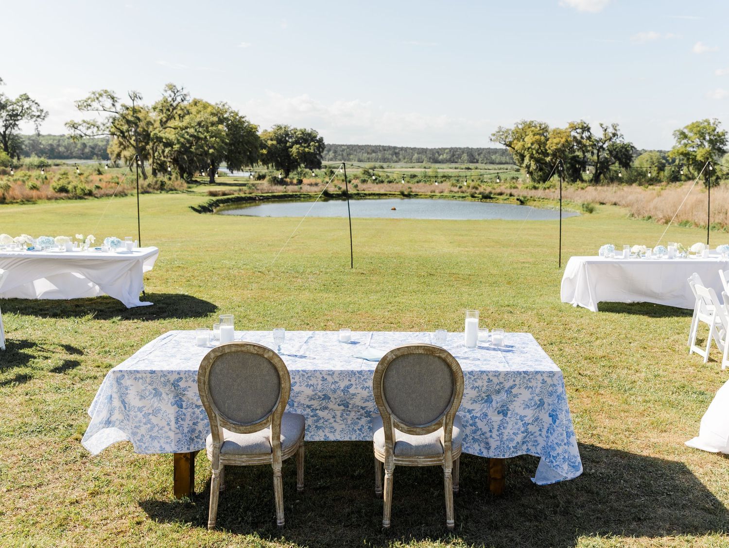 Outdoor wedding reception tables with blue floral linens set on green lawn overlooking serene pond.