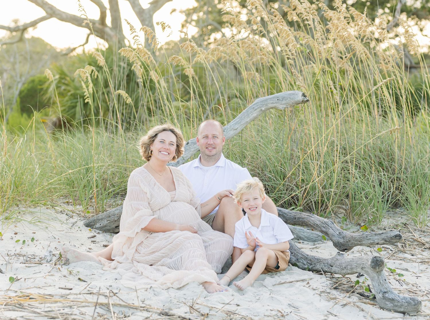 Family maternity photo on beach with tall grass and natural coastal backdrop.