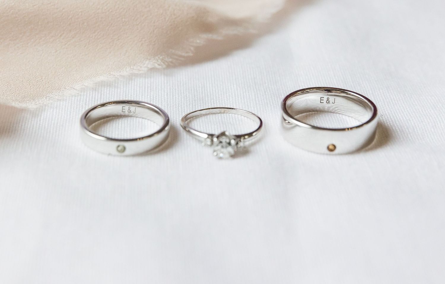 Three elegant silver rings displayed on white fabric, including a solitaire engagement ring and two plain bands.
