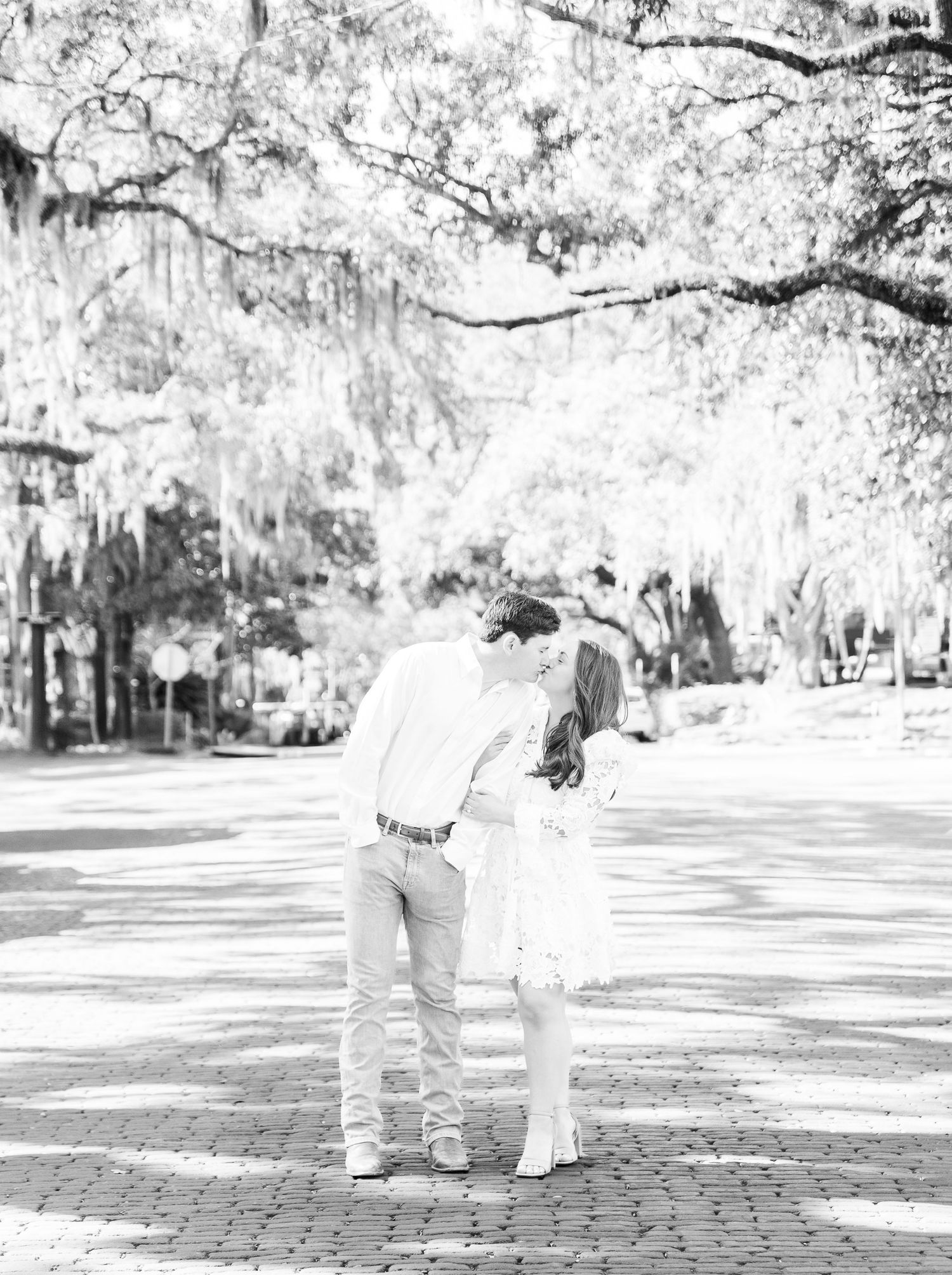 A romantic black and white photo of a couple walking under Spanish moss draped trees on a brick path.