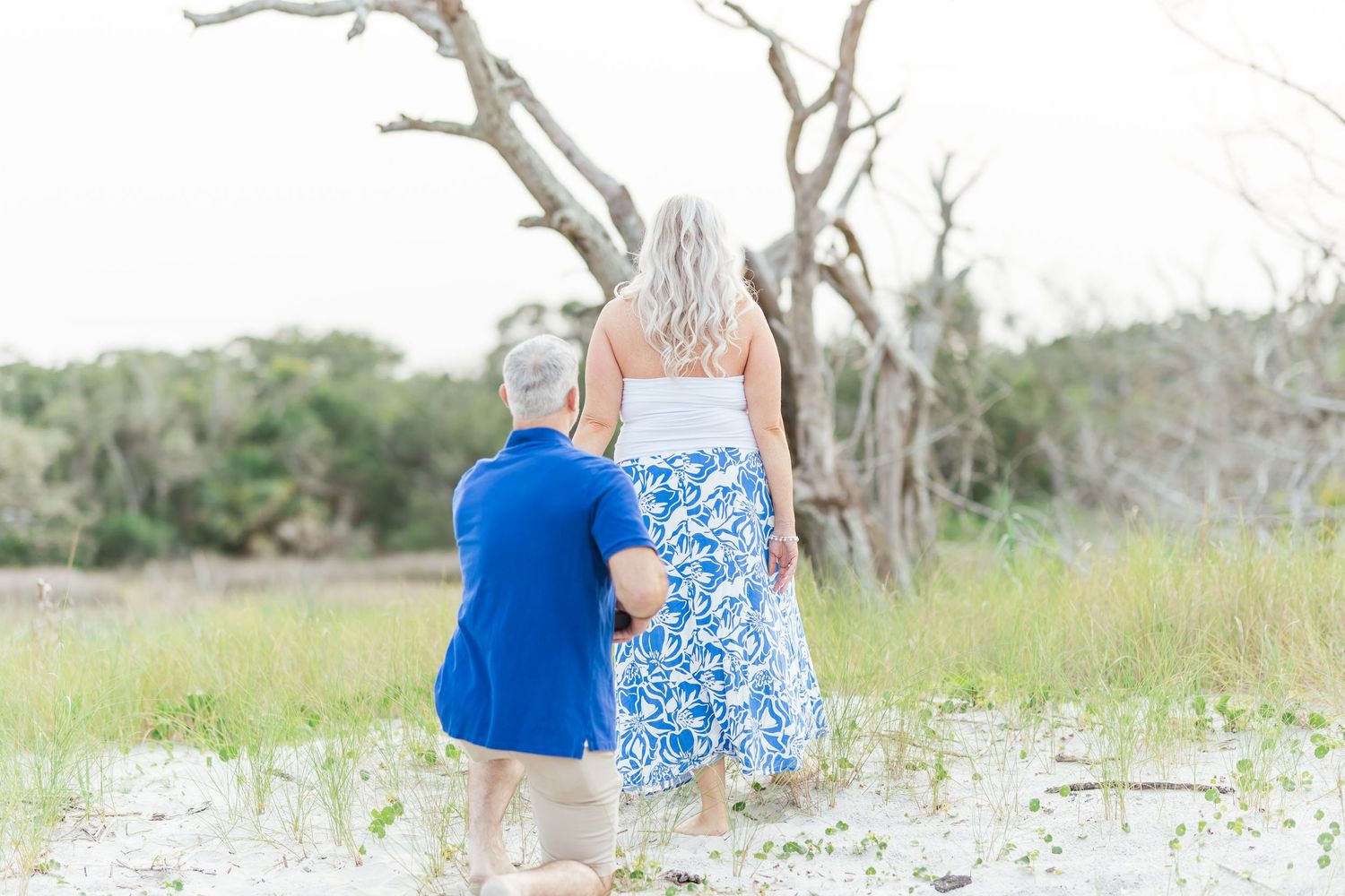 Two people stand together on a beach with bare trees, wearing blue clothes against a coastal landscape.