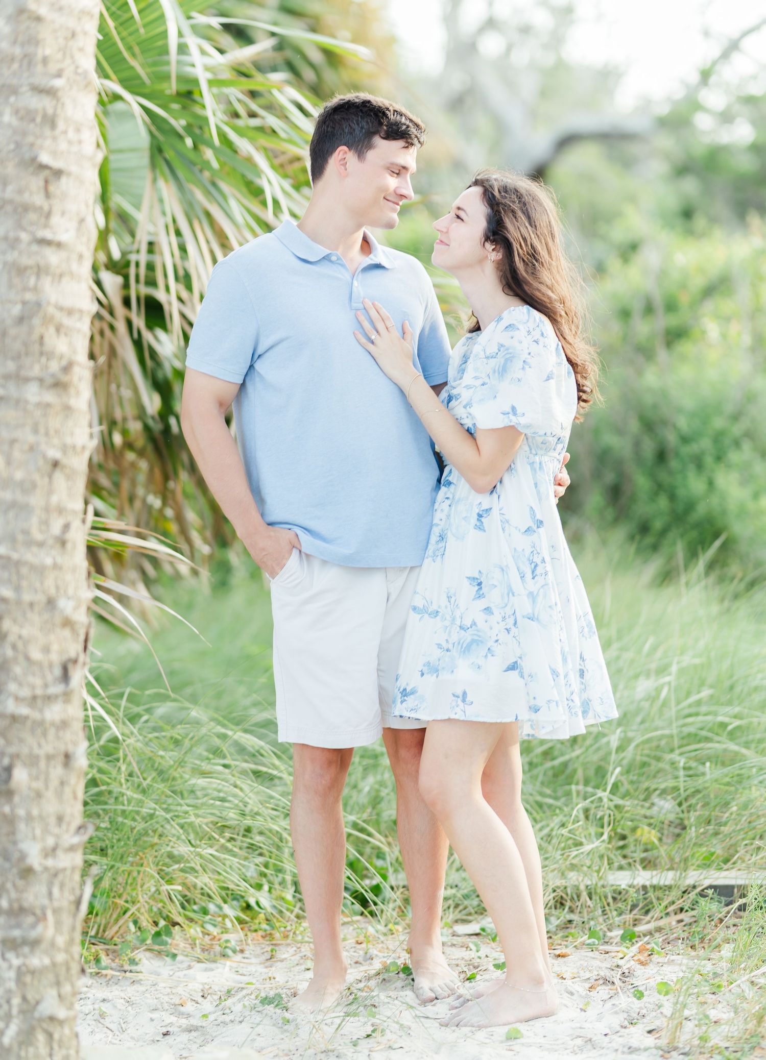 Couple in matching blue outfits pose together on sandy path with tropical foliage in background.
