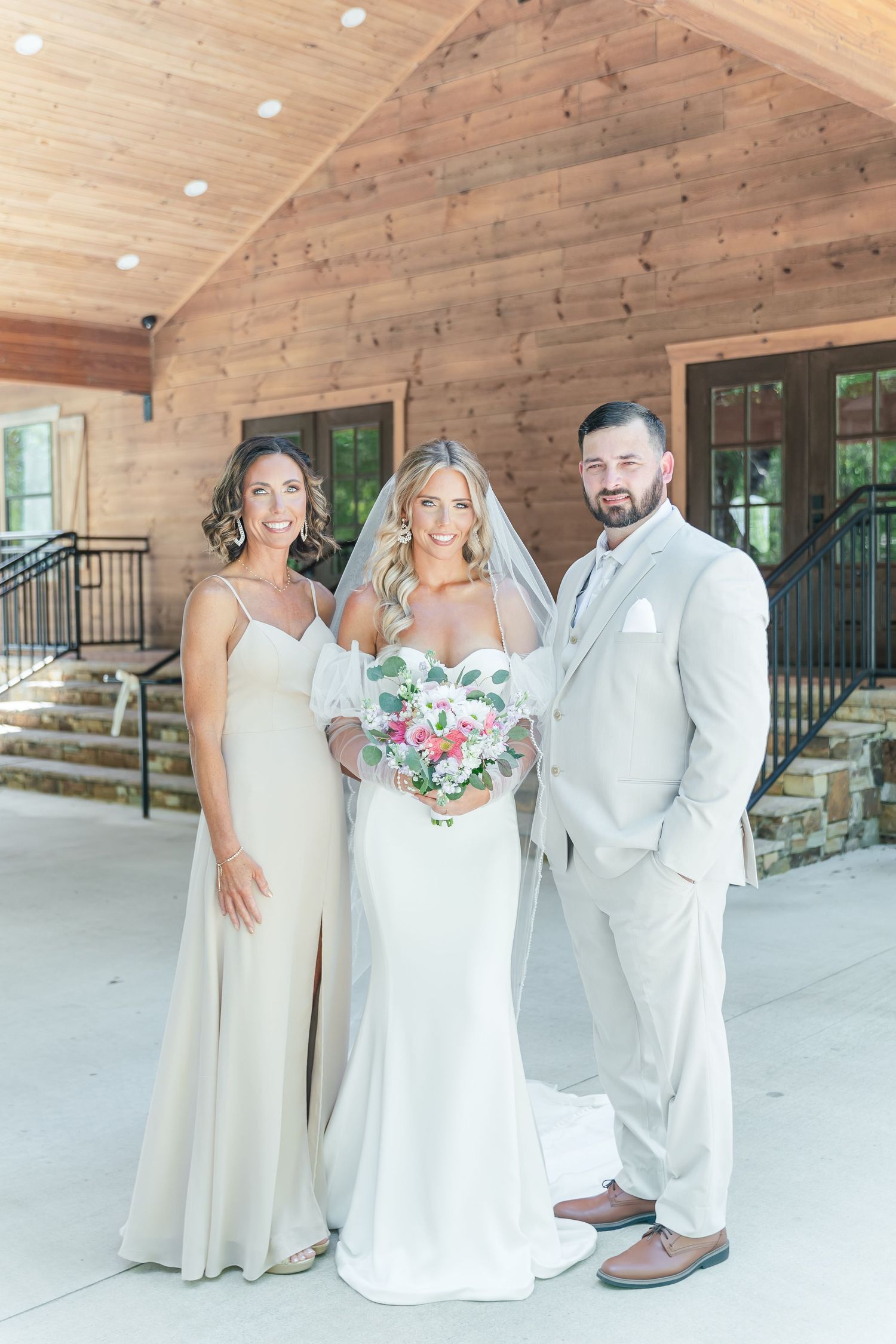 Wedding party poses in elegant white attire against rustic wooden backdrop with floral bouquet.
