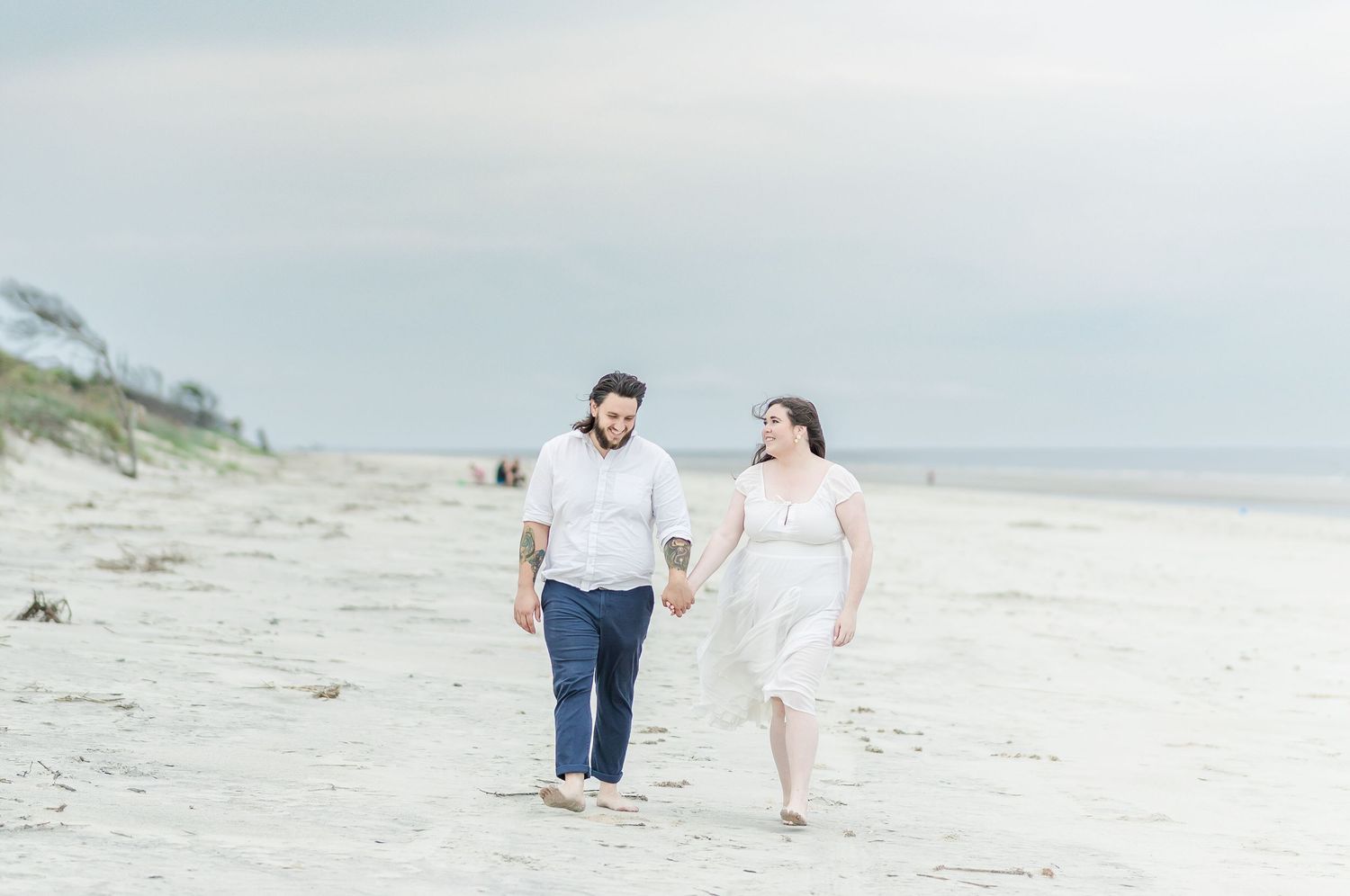 Couple walking hand in hand along a serene beach on an overcast day.