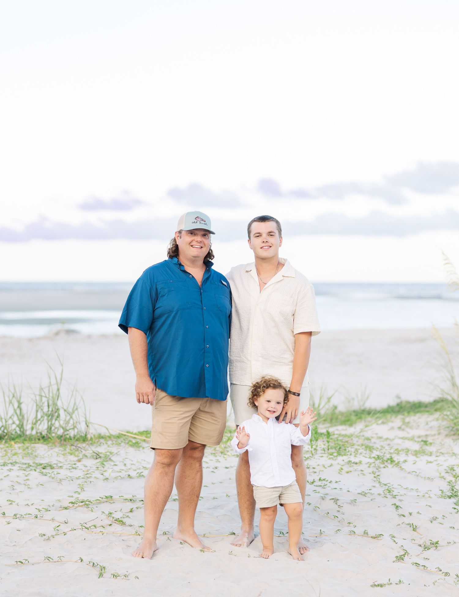 A family group stands together on a sandy beach with beach grass and ocean waves visible in the background.