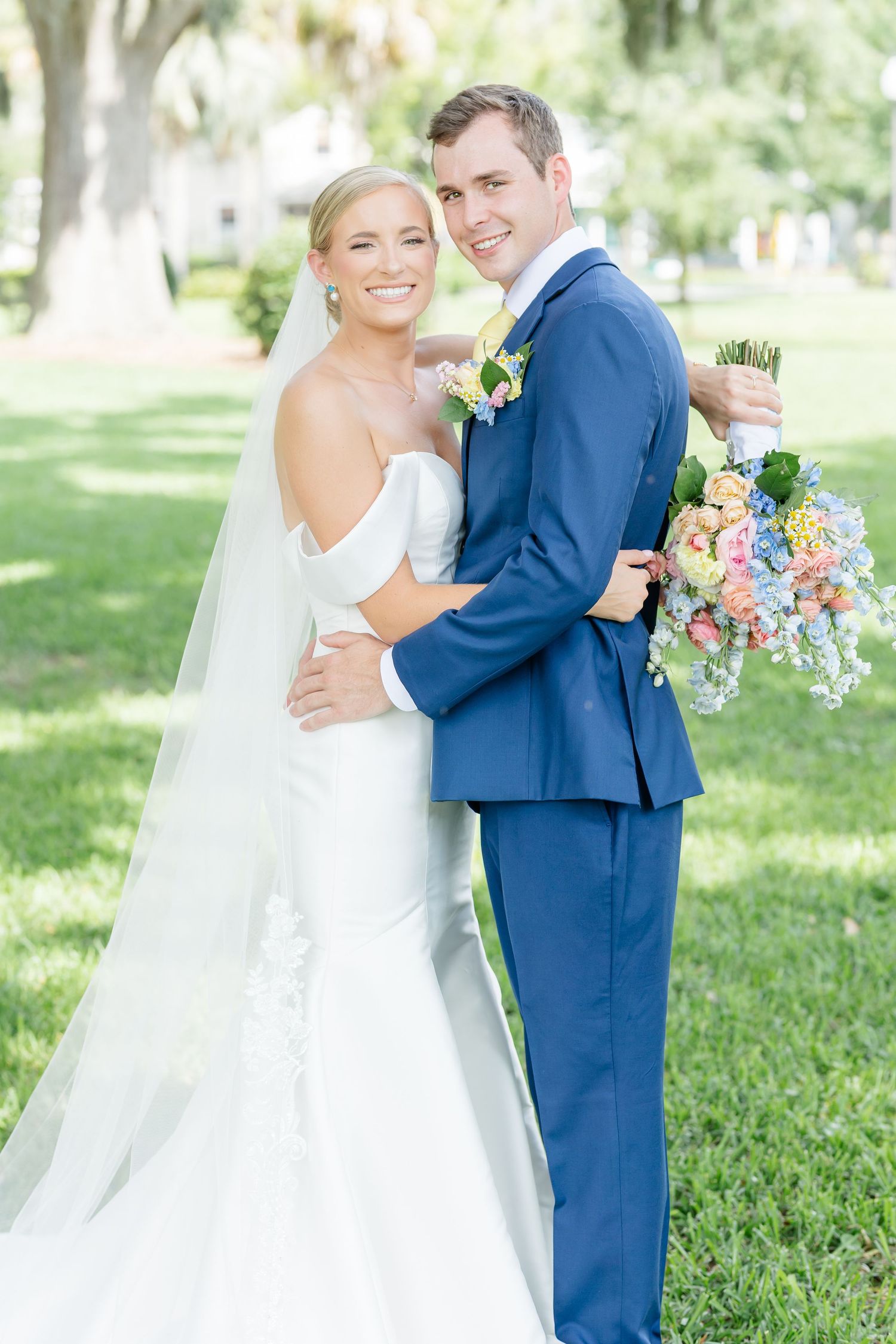 A newlywed couple poses outdoors with the bride in white gown and groom in navy suit holding a colorful floral bouquet.