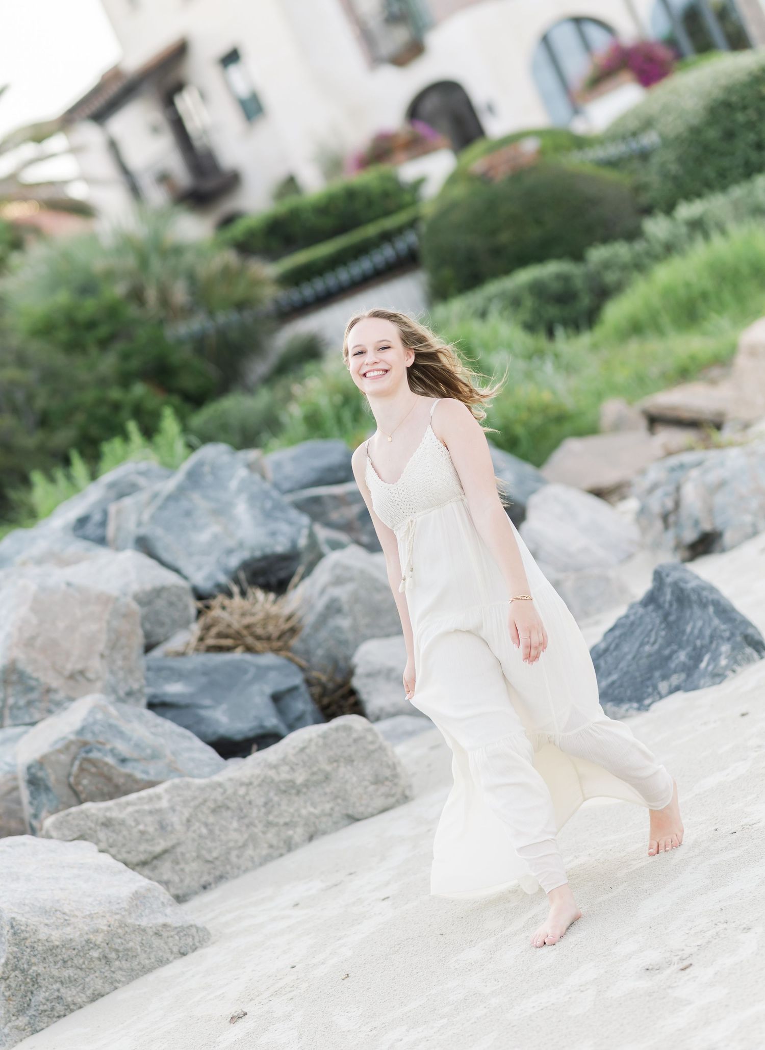 White flowing dress catches the breeze along rocky coastal shoreline with white building in background.