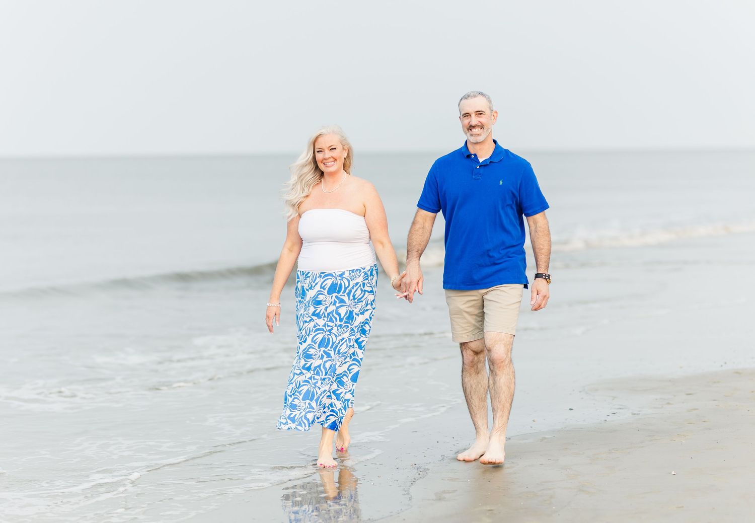 A couple walks hand in hand along a misty beach shore, wearing blue and white coordinated outfits.