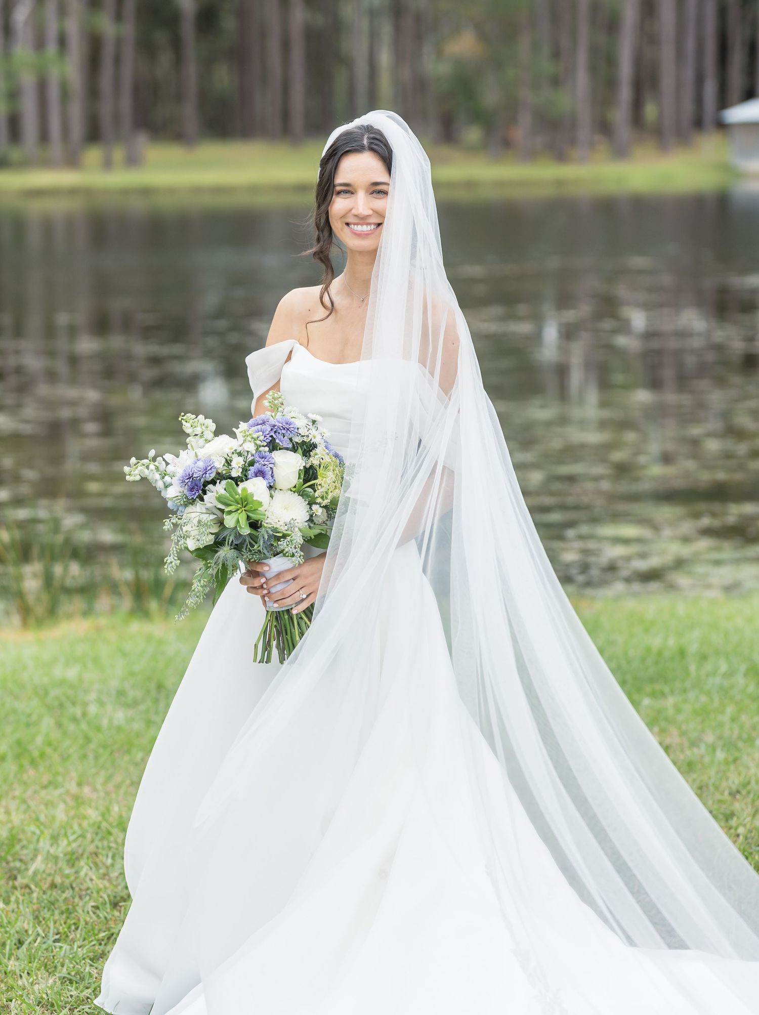 A bride in a flowing white gown and veil holds a purple and white bouquet by a lakeside.