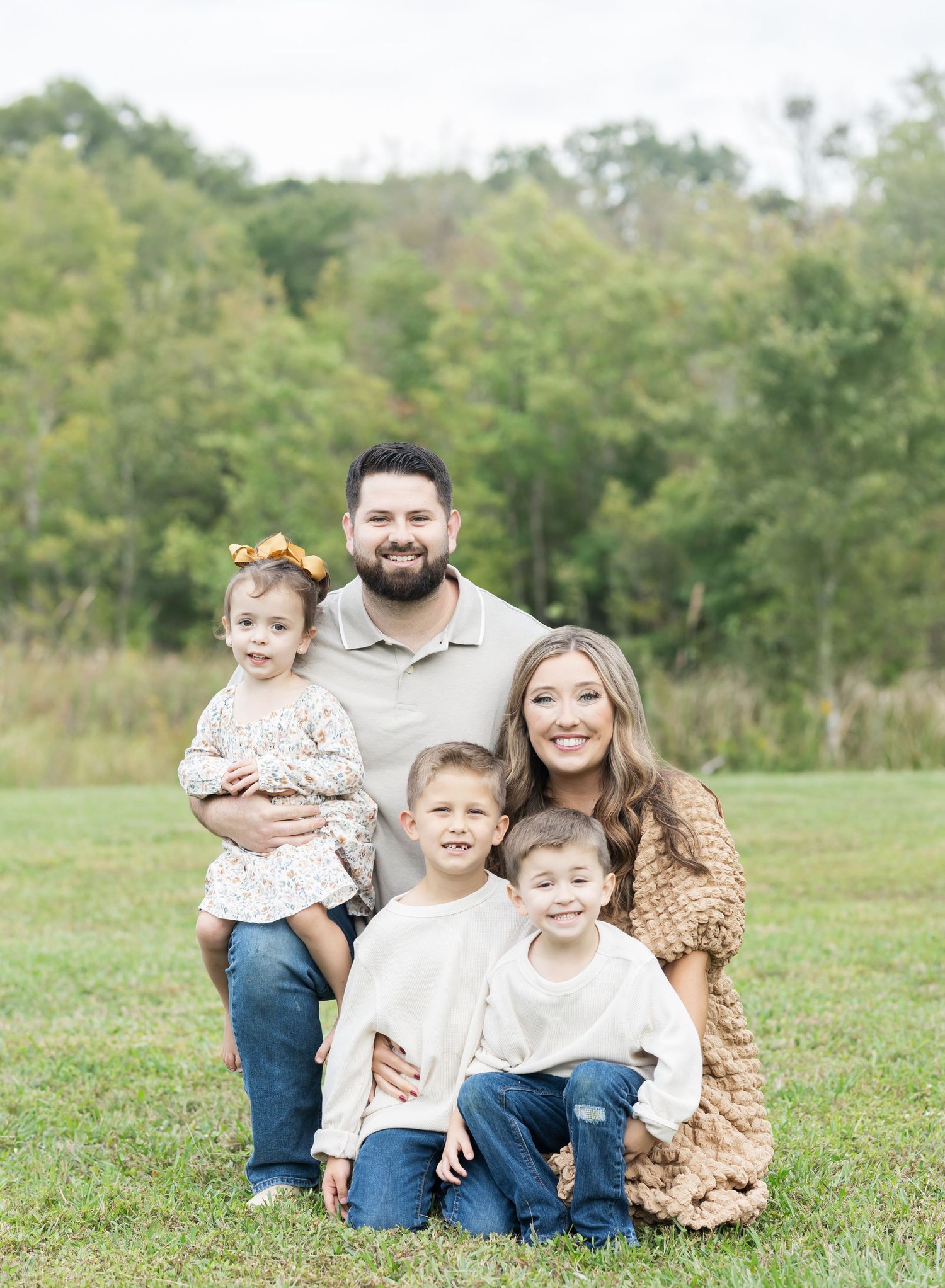 A family group portrait taken outdoors against a backdrop of green trees and grass.