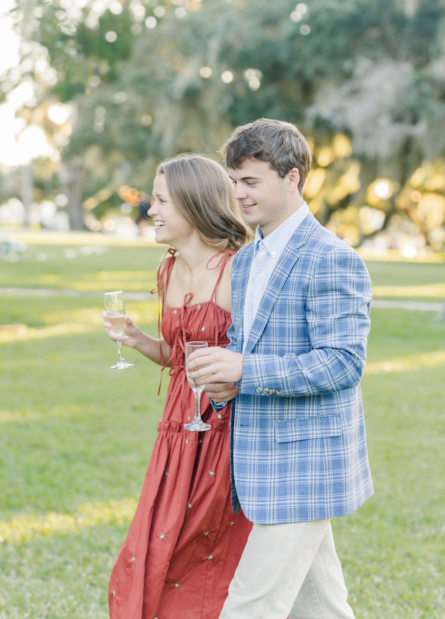 A couple in a park sharing champagne, with her in a rust-colored dress and him in a blue plaid blazer.