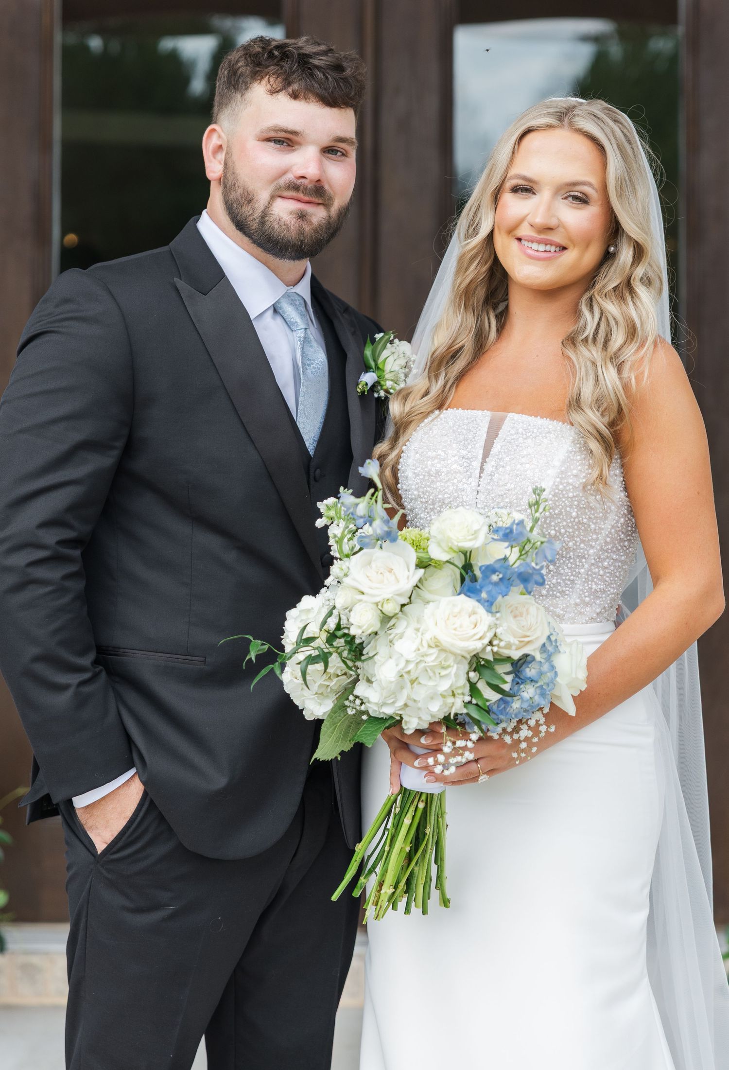 Bride holding white and blue bouquet stands with groom in black suit at wedding venue.