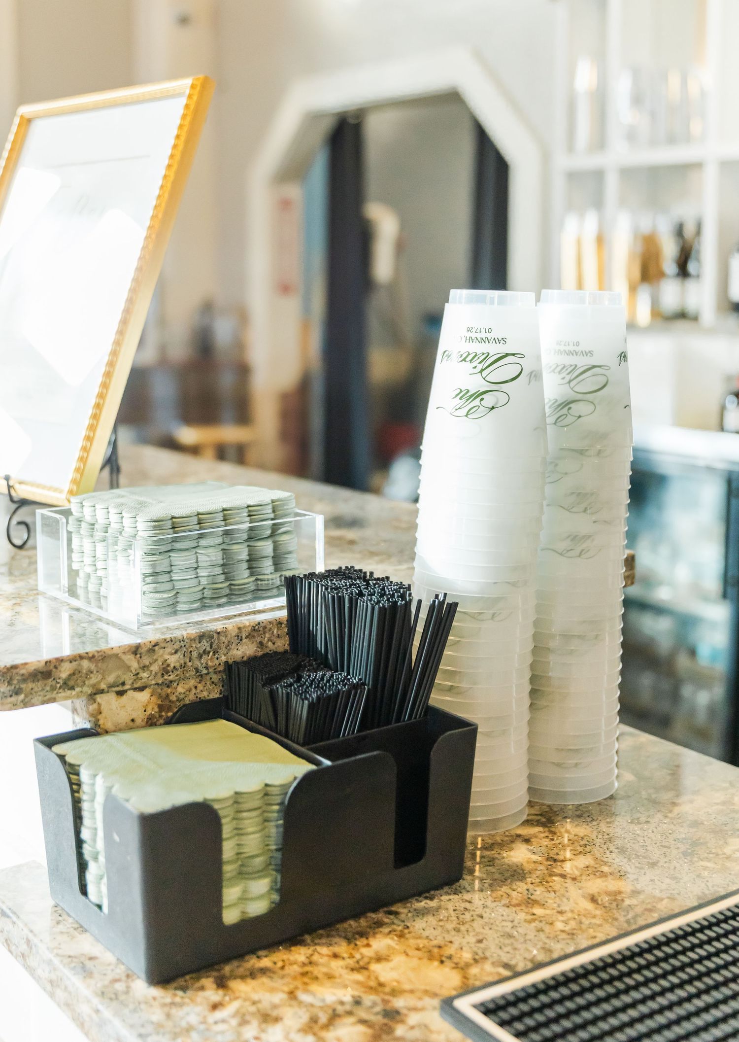 Coffee bar setup with stacked branded cups, black straw holder, napkin dispenser, and gold-framed sign on marble counter.
