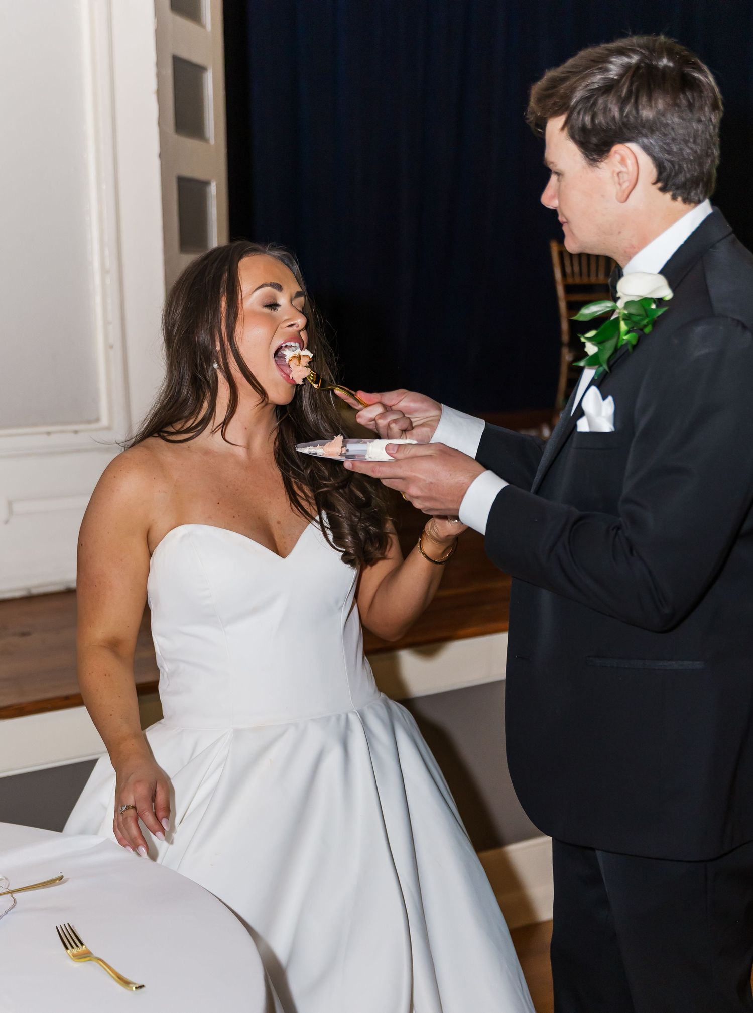 Bride laughing as groom feeds her wedding cake at reception, wearing strapless white gown and black tuxedo.