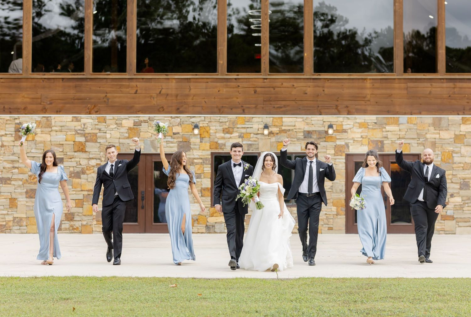 Wedding party in formal attire walks and poses playfully along a brick wall outdoors during a celebration.