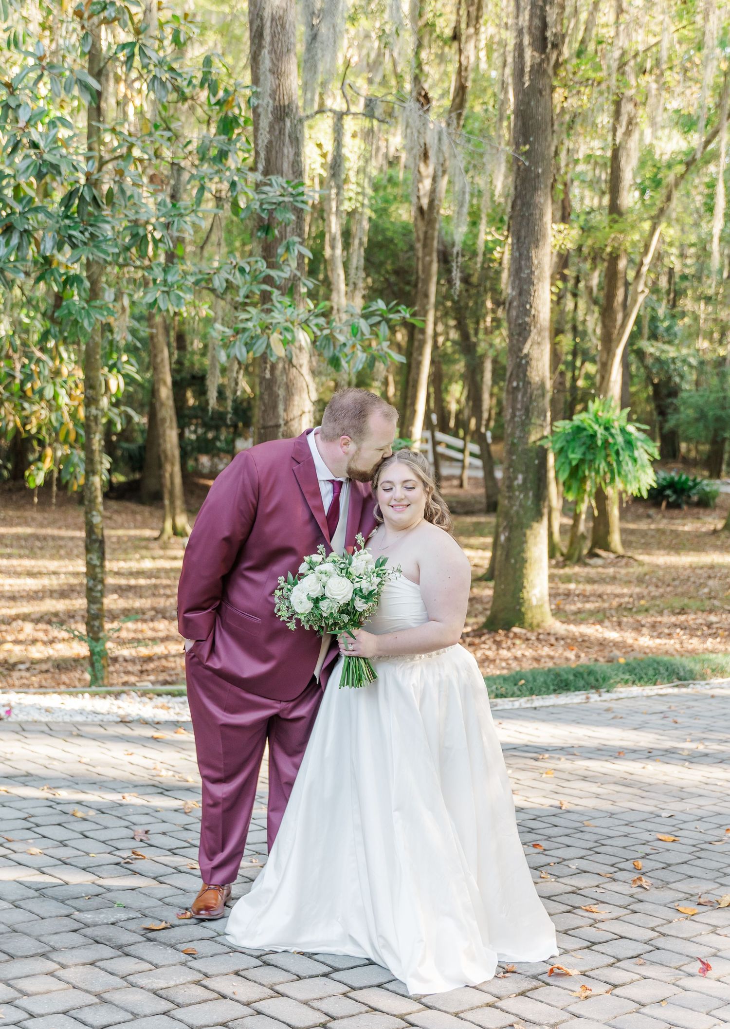 Wedding couple in burgundy and white attire posing together on a brick pathway surrounded by trees.