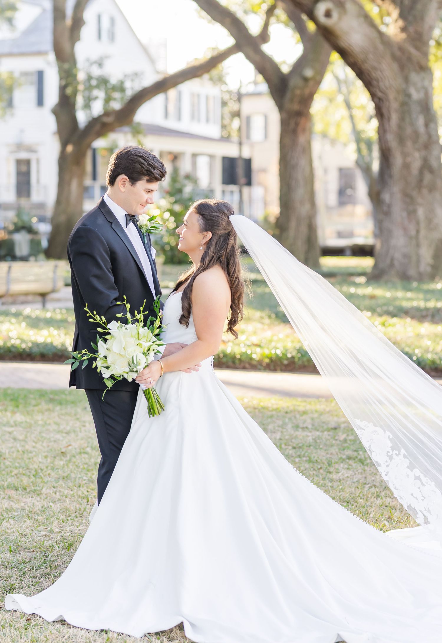 A romantic wedding photo in a sunlit park with autumn trees, featuring a flowing white gown and long veil.