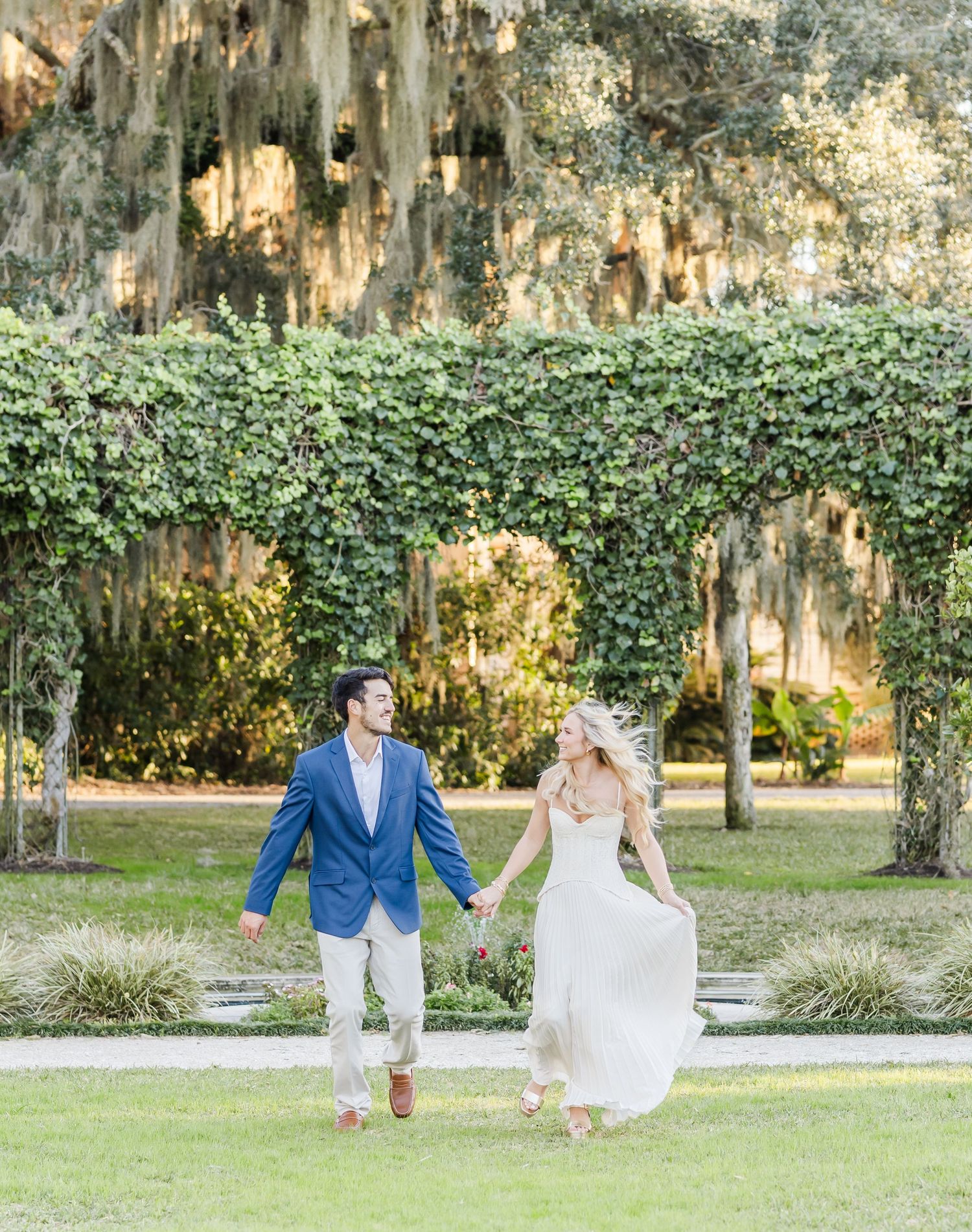 Newlywed couple playfully walking and running through a garden with Spanish moss hanging from trees.