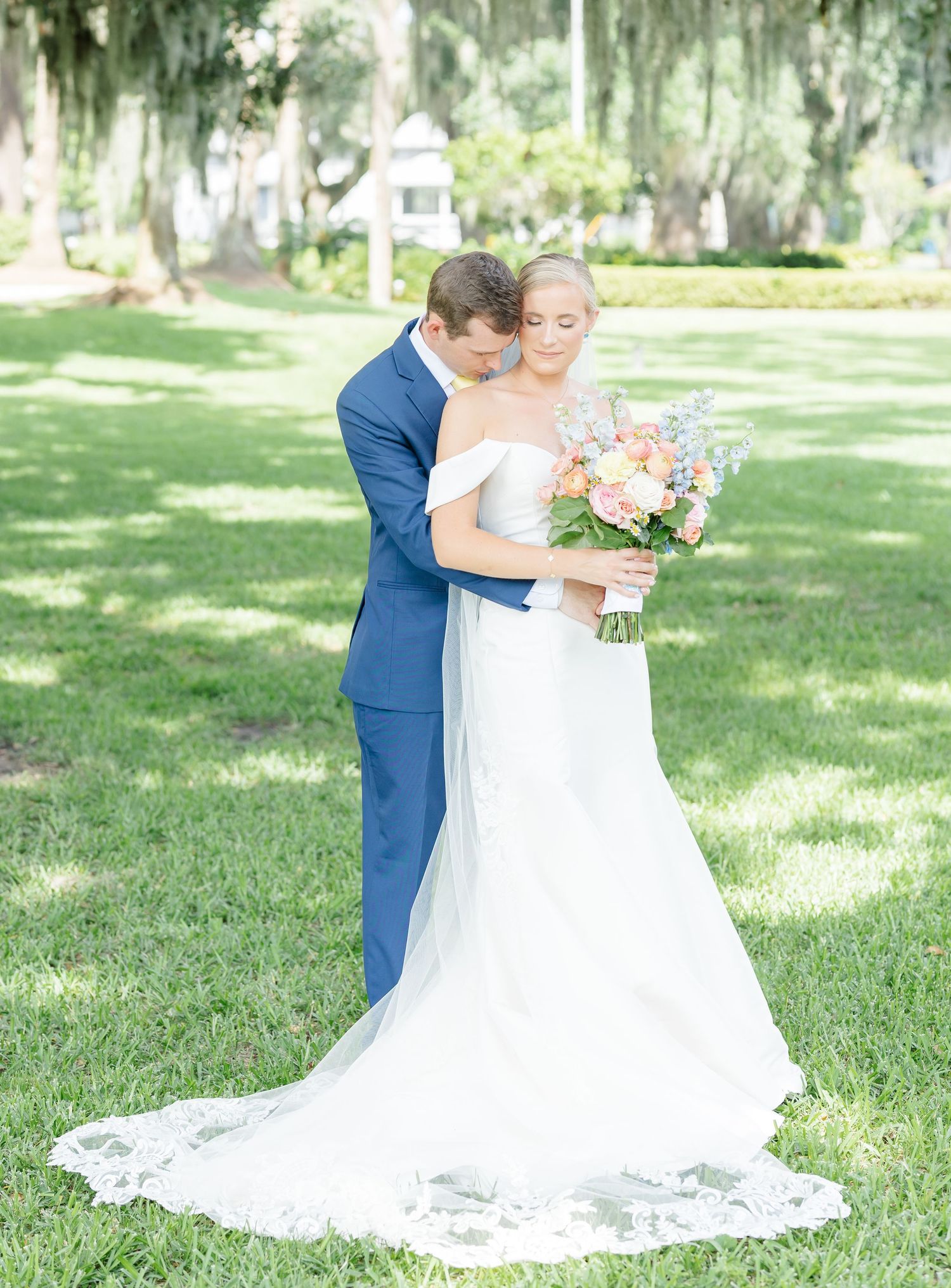 A couple shares an intimate first look moment in a sunlit garden with Spanish moss draped trees in the background.