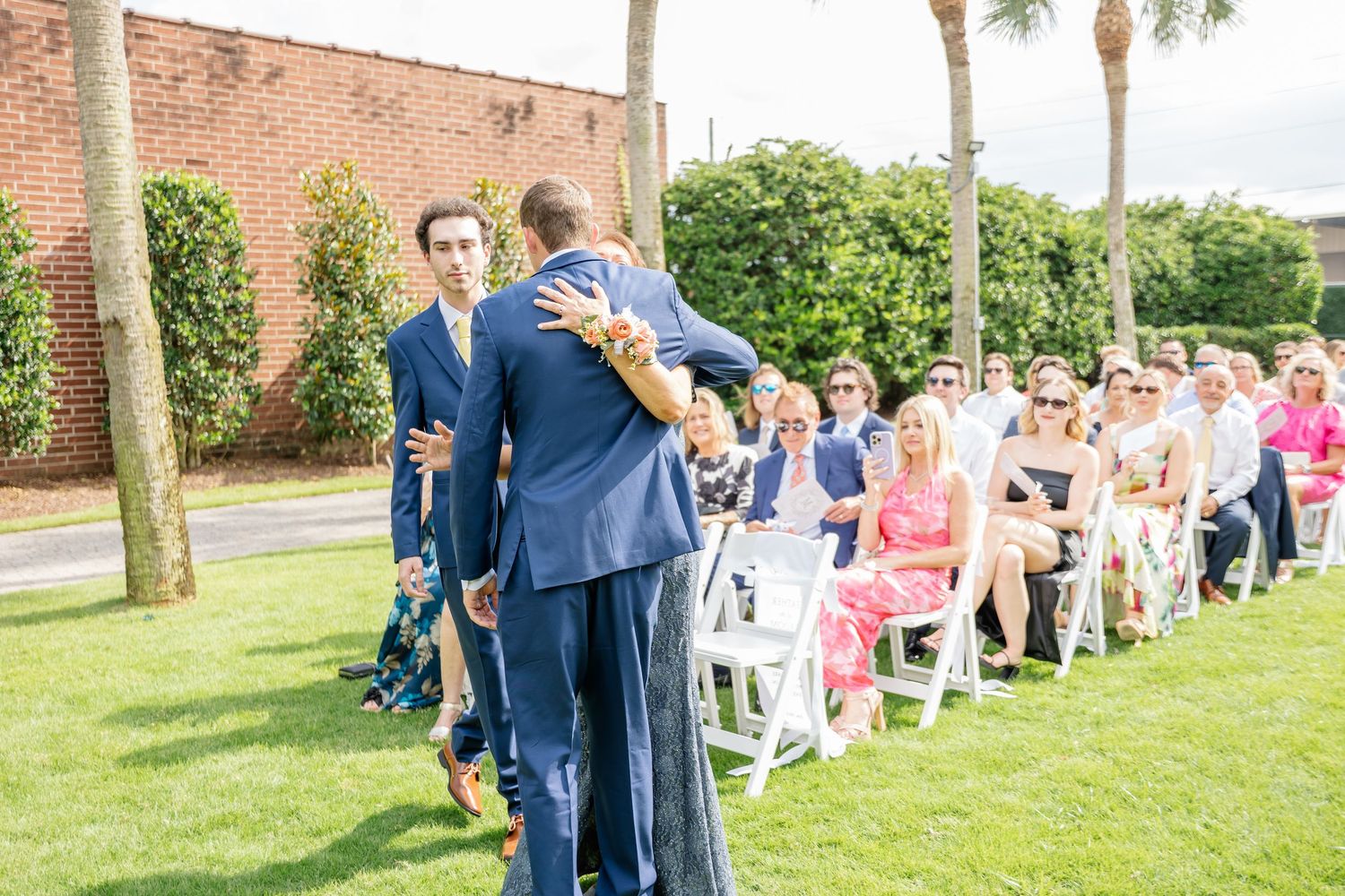 Wedding guests stand in a receiving line on a grassy lawn with palm trees and a brick building in the background.