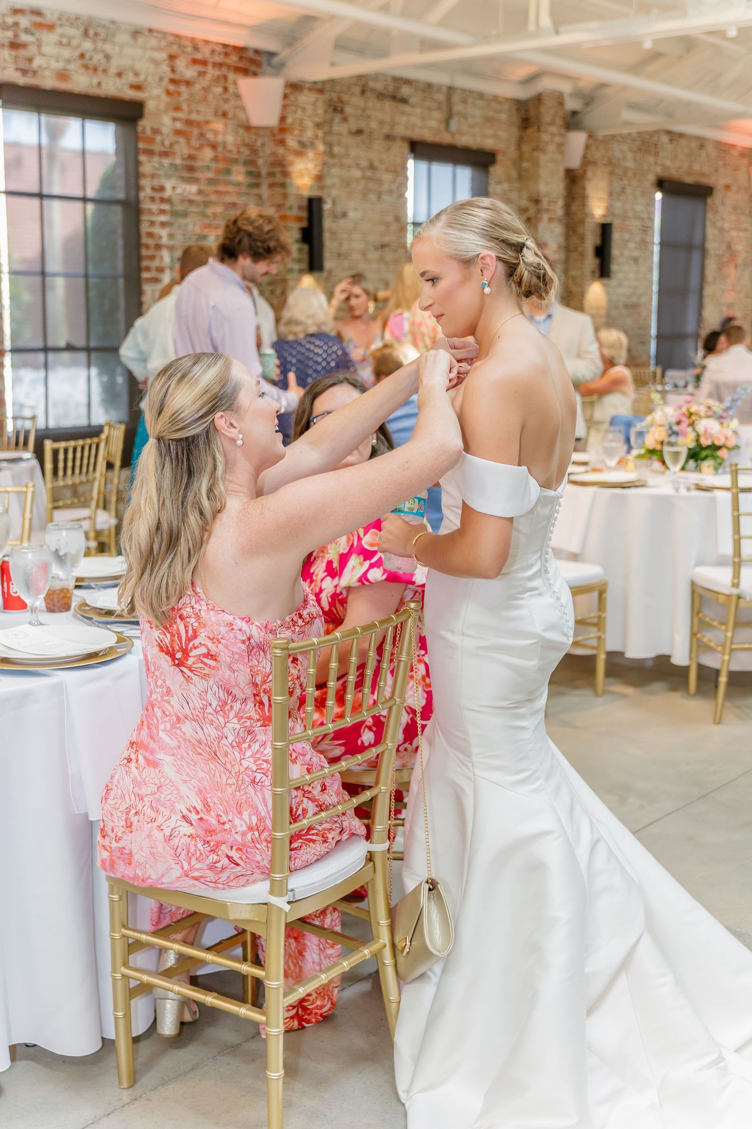 Wedding guests interact during an elegant reception in a rustic brick venue with gold chairs and white tablecloths.