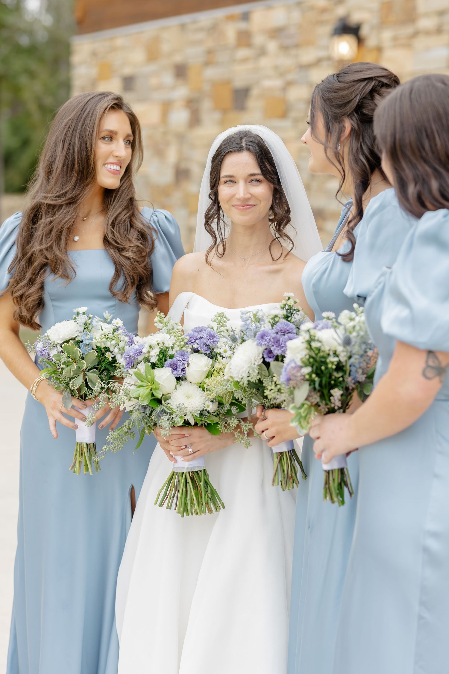 A bride in white gown stands with bridesmaids in light blue dresses holding purple and white floral bouquets.