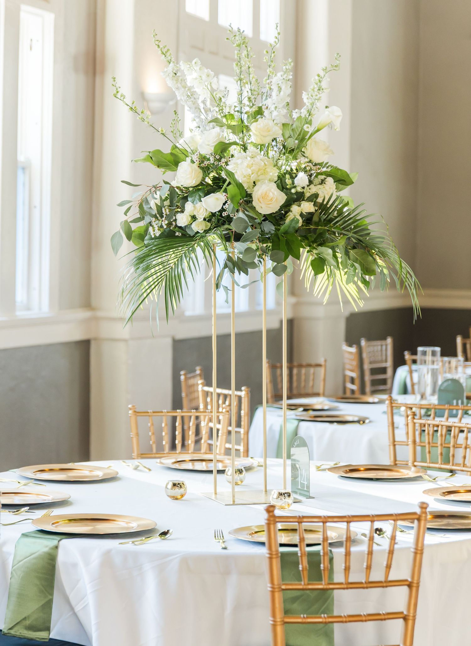 Elegant wedding reception hall with white linens, gold chairs, and elevated floral arrangements.
