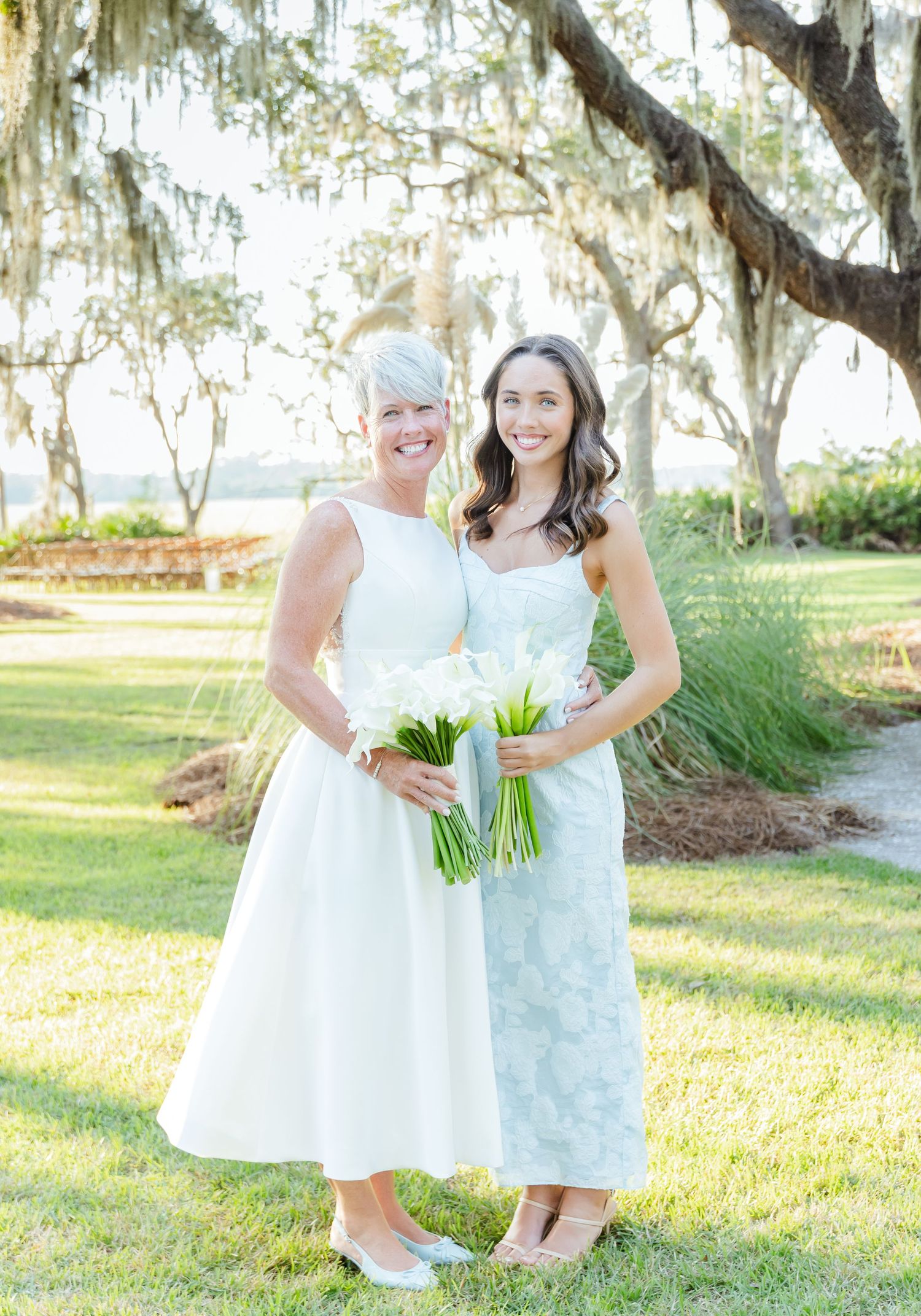 Bridesmaids in white dresses holding bouquets pose together on a sunny lawn with moss-covered trees in background.