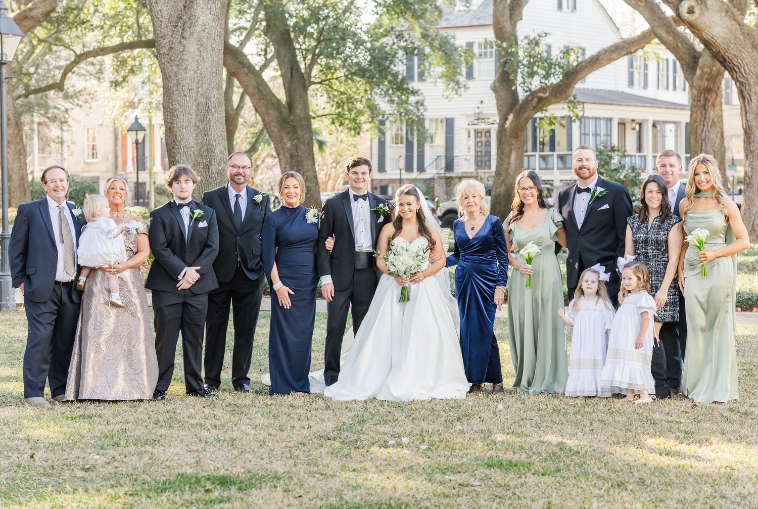 Large wedding party group photo with groomsmen and bridesmaids on a sunny outdoor lawn.