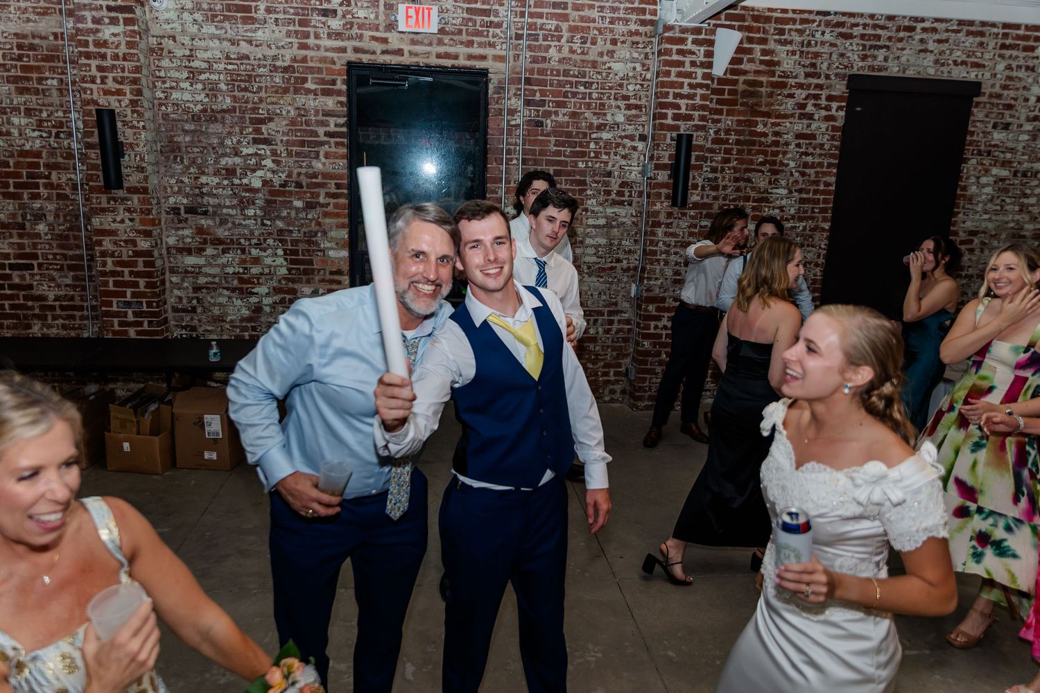 Wedding guests dance and celebrate together on a rustic brick-walled dance floor at an indoor reception venue.
