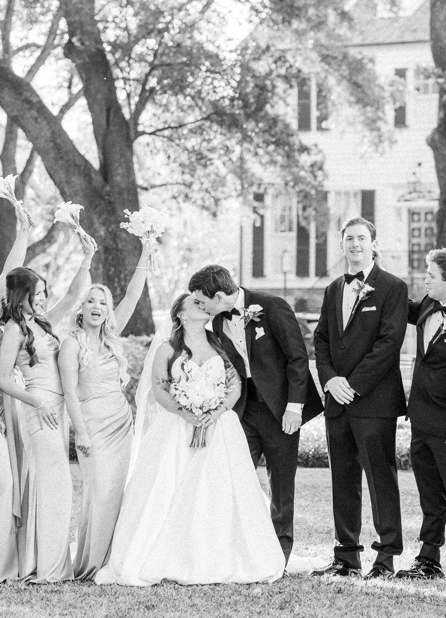 Wedding party in formal attire poses playfully while lying on grass in black and white photograph.