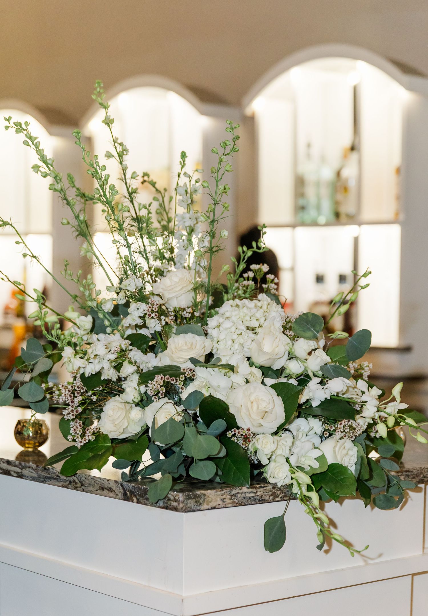 Elegant white floral centerpiece with roses, eucalyptus leaves and delicate greenery on white pedestal.