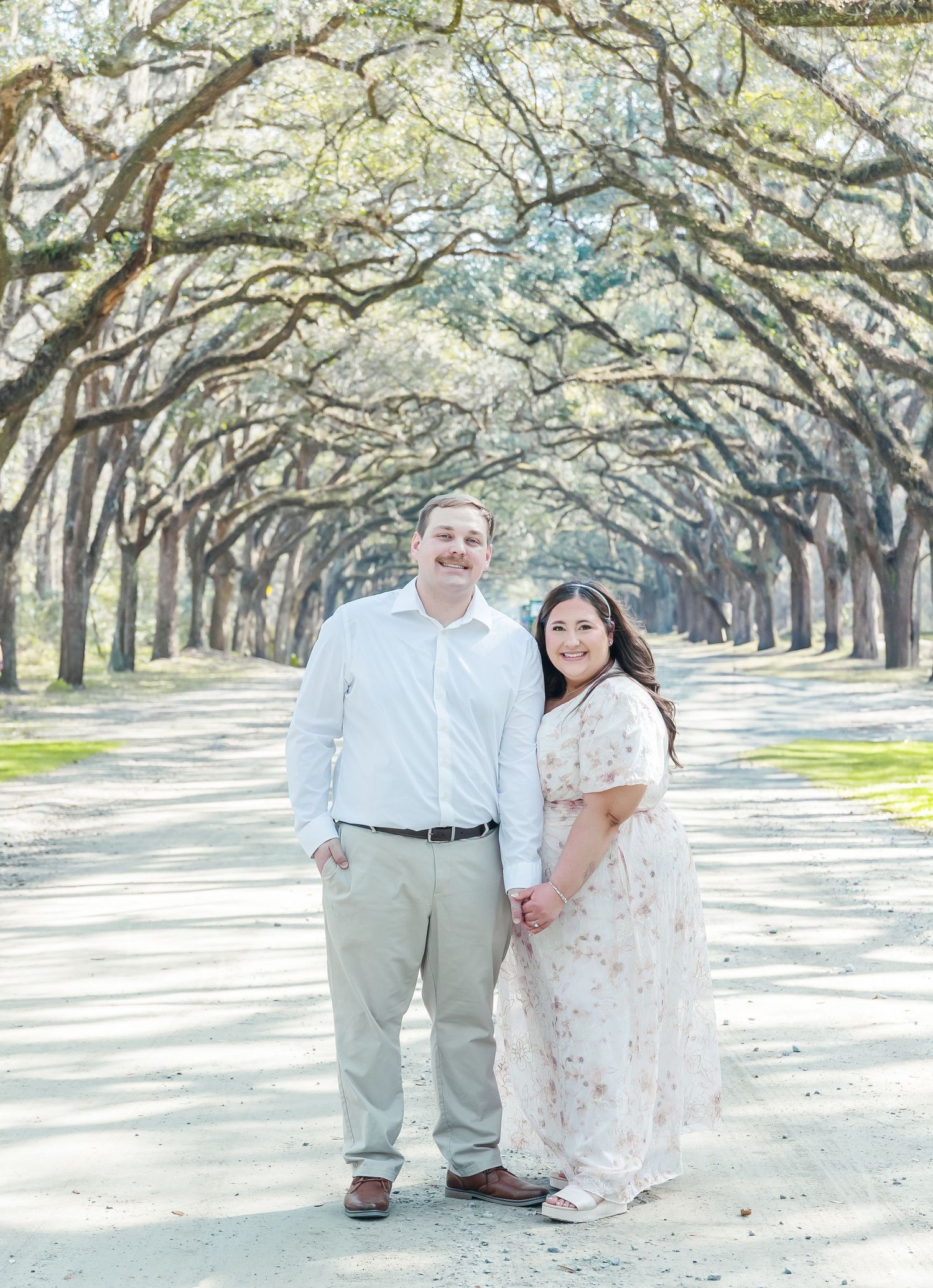 Engaged couple standing on a path lined with oak trees forming a natural tunnel overhead.